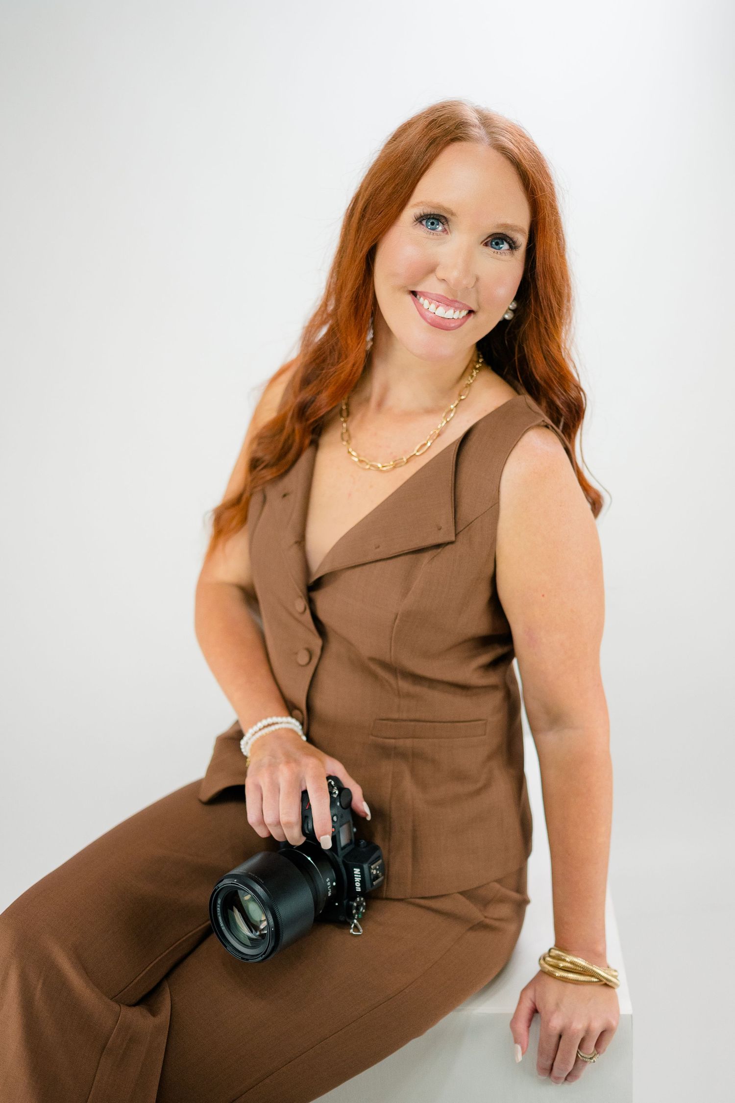Red-haired woman in brown jumpsuit holding a camera, smiling confidently in a professional studio setting.