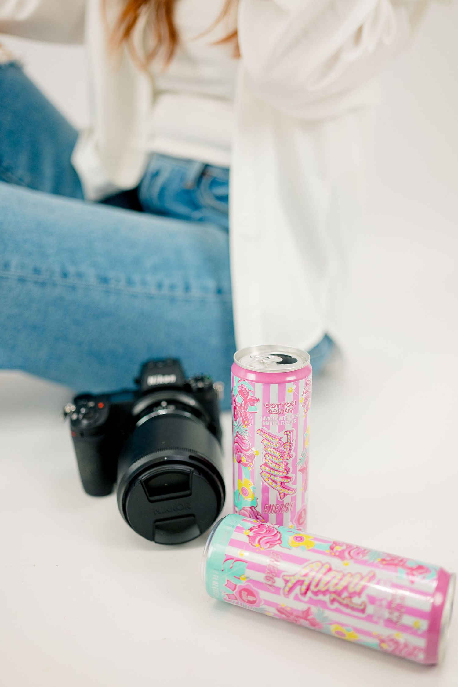 Pink Alani Nu energy drink cans beside a Nikon camera, with a woman in jeans and white top sitting nearby.