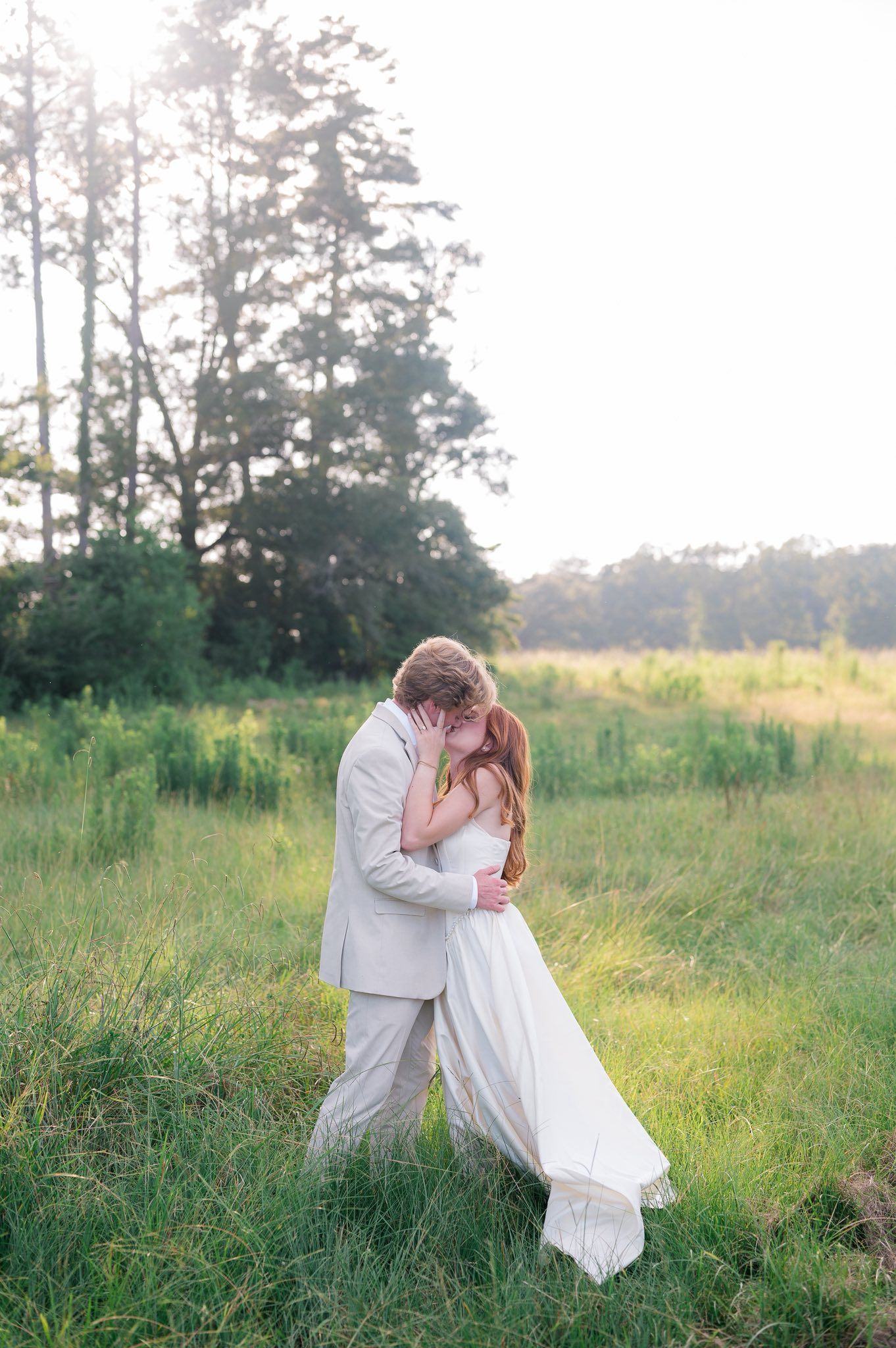 A romantic couple in white attire shares an intimate moment in a sunlit meadow with tall grass and trees in the background.