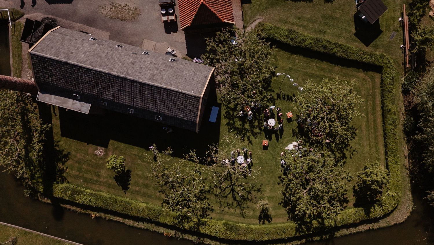 Aerial view of a rural farmhouse with a gray roof surrounded by lush green grass and trees with a group of people gathered outside.