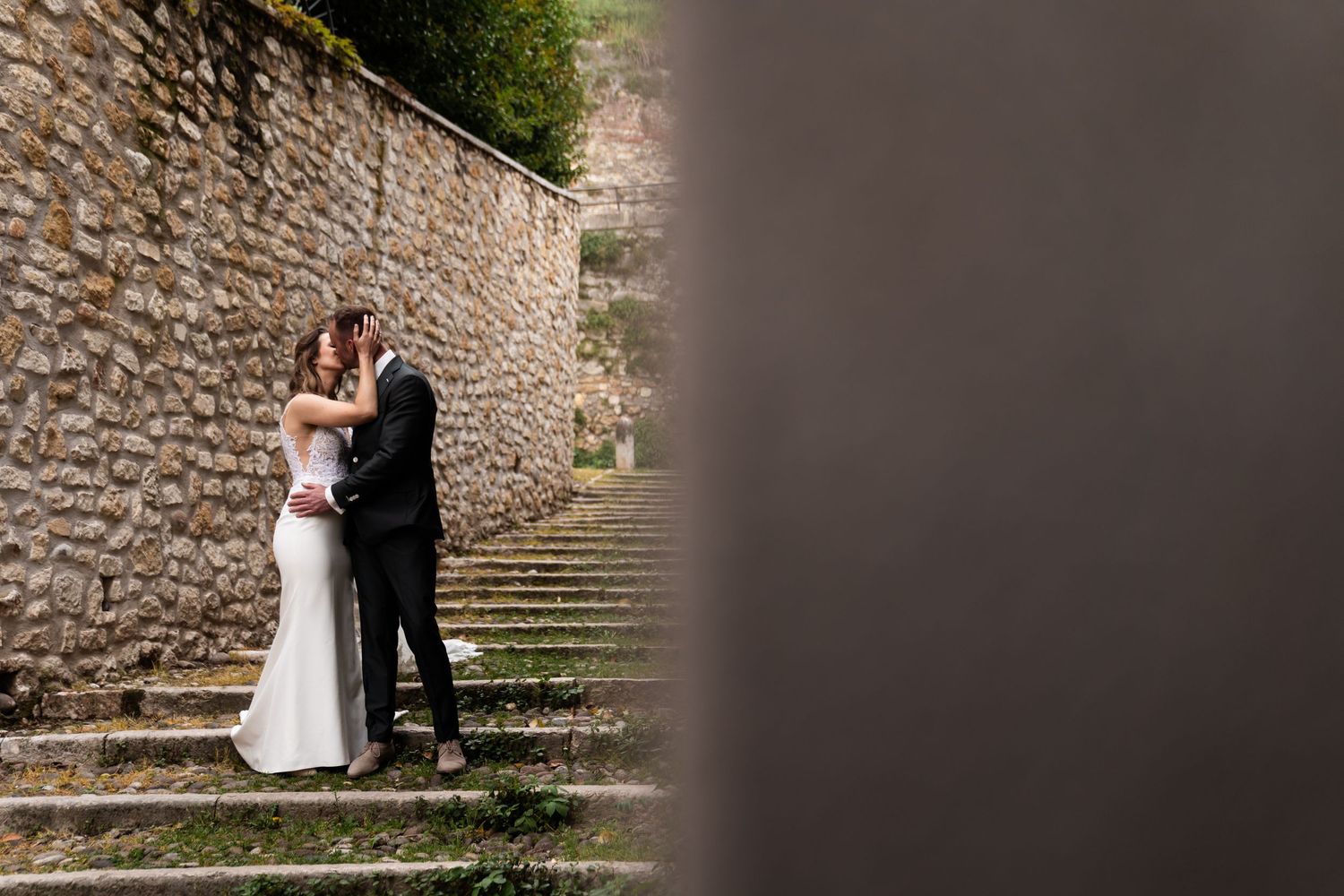 A couple in formal wedding attire embraces on stone steps against an ancient stone wall.