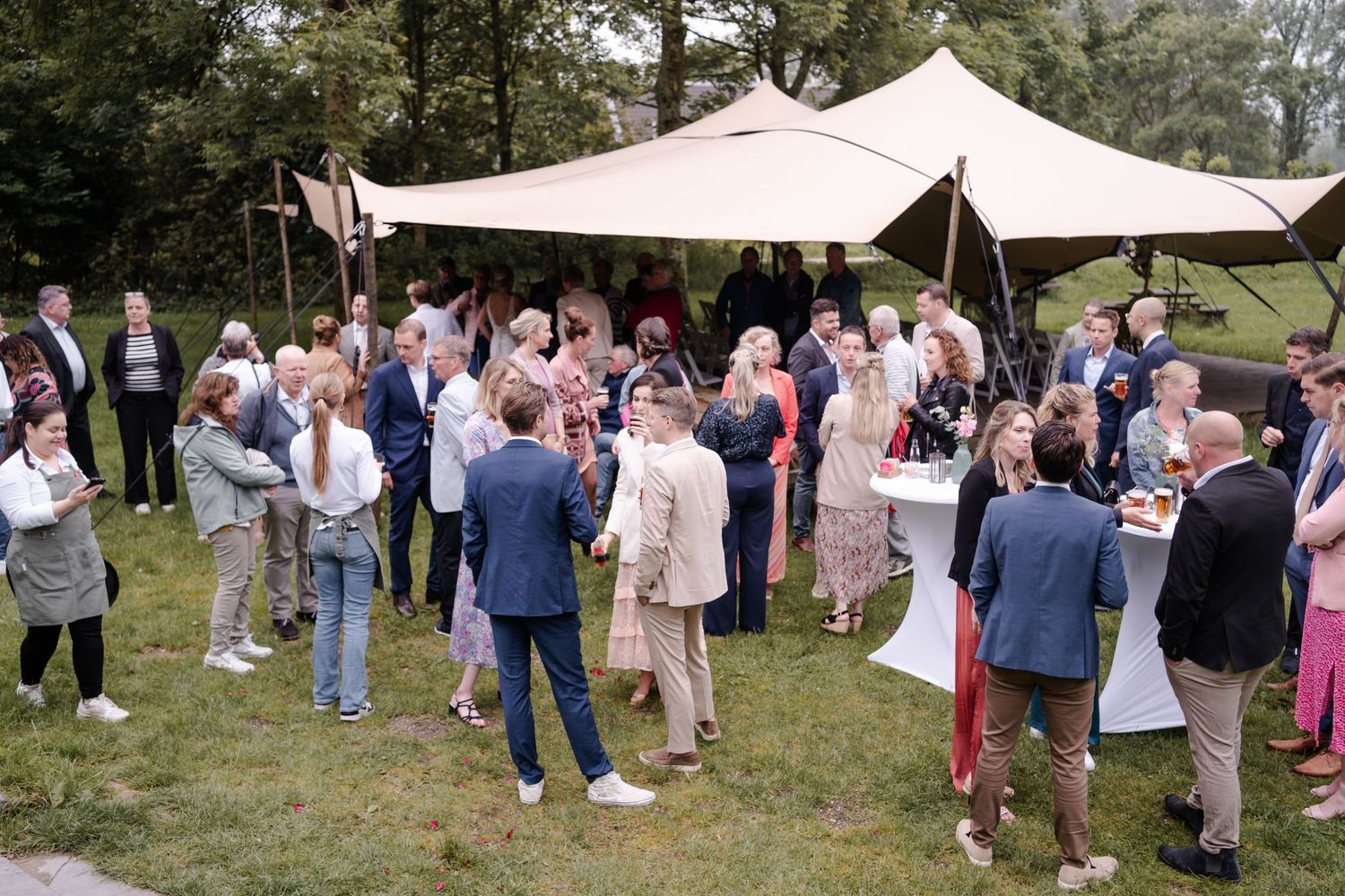 A group of wedding guests gather under a beige canopy tent during an outdoor garden reception.