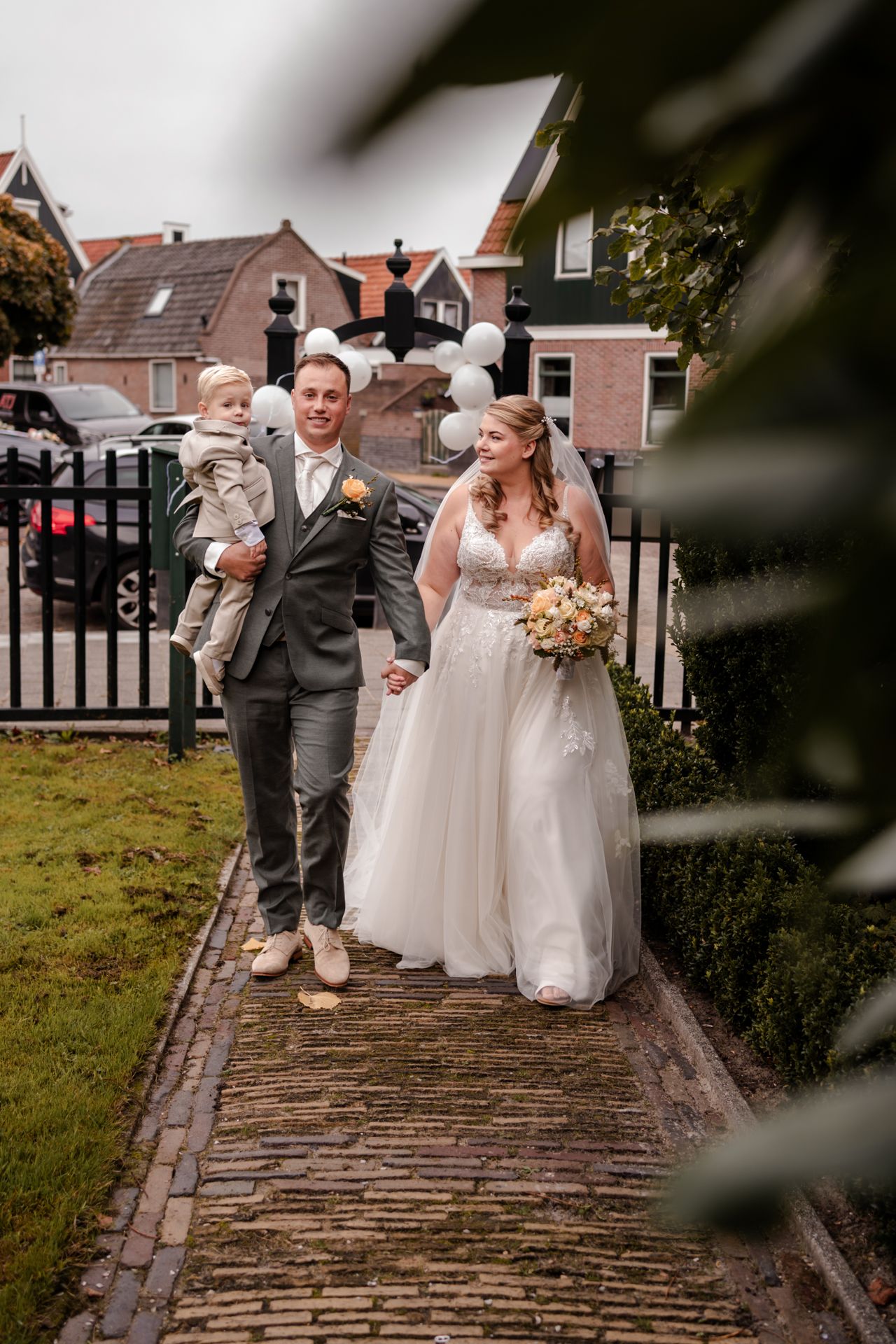 Wedding couple walks down brick path with child while carrying floral bouquet.