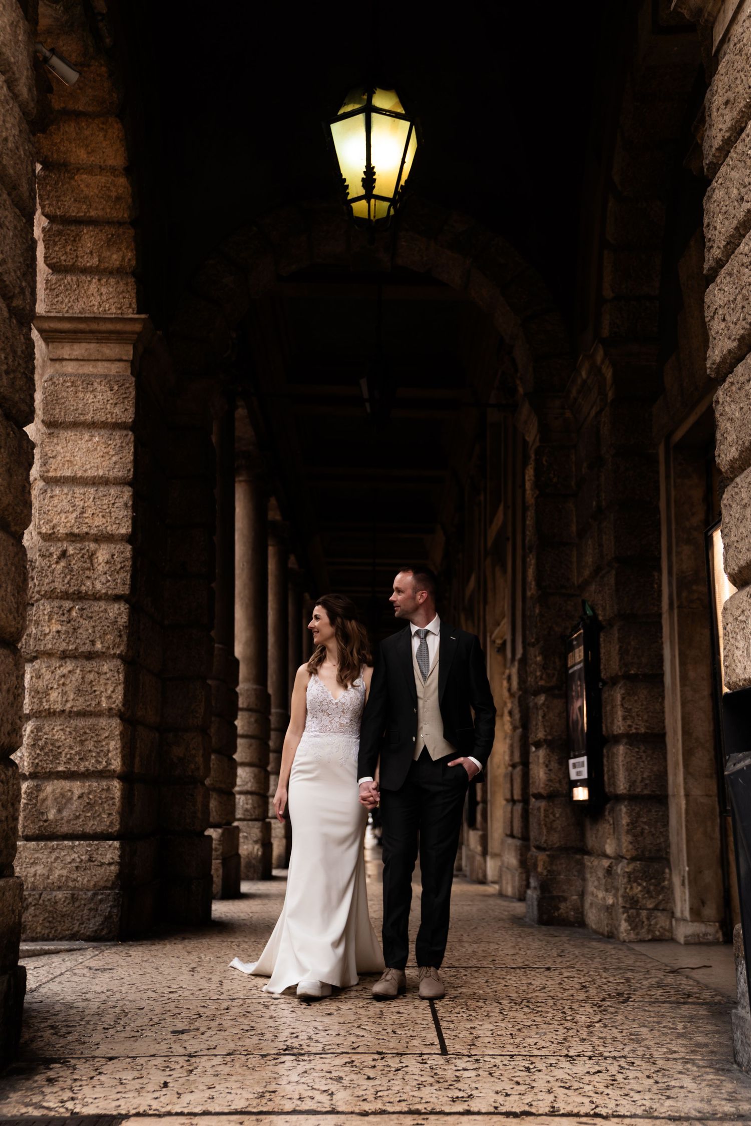 A couple stands in a romantic stone archway corridor with glowing lanterns overhead in an elegant European setting.