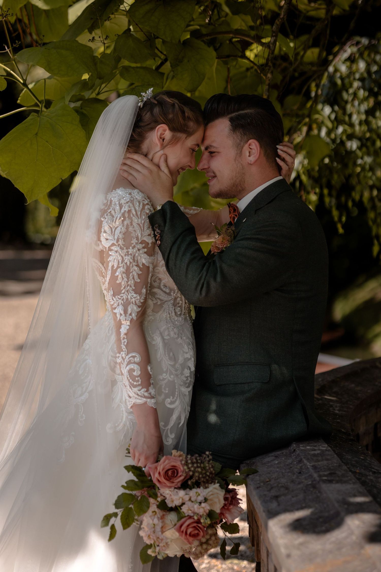 A romantic wedding couple embraces under green foliage in an intimate outdoor setting.