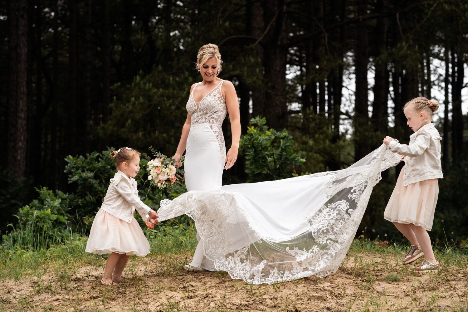 A lace wedding dress train is playfully held by two young children in white outfits against a wooded forest backdrop.