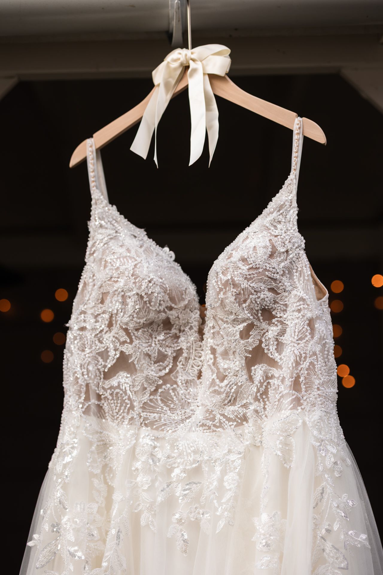 A delicate white lace wedding dress with intricate floral details hangs on a wooden hanger with a white bow.