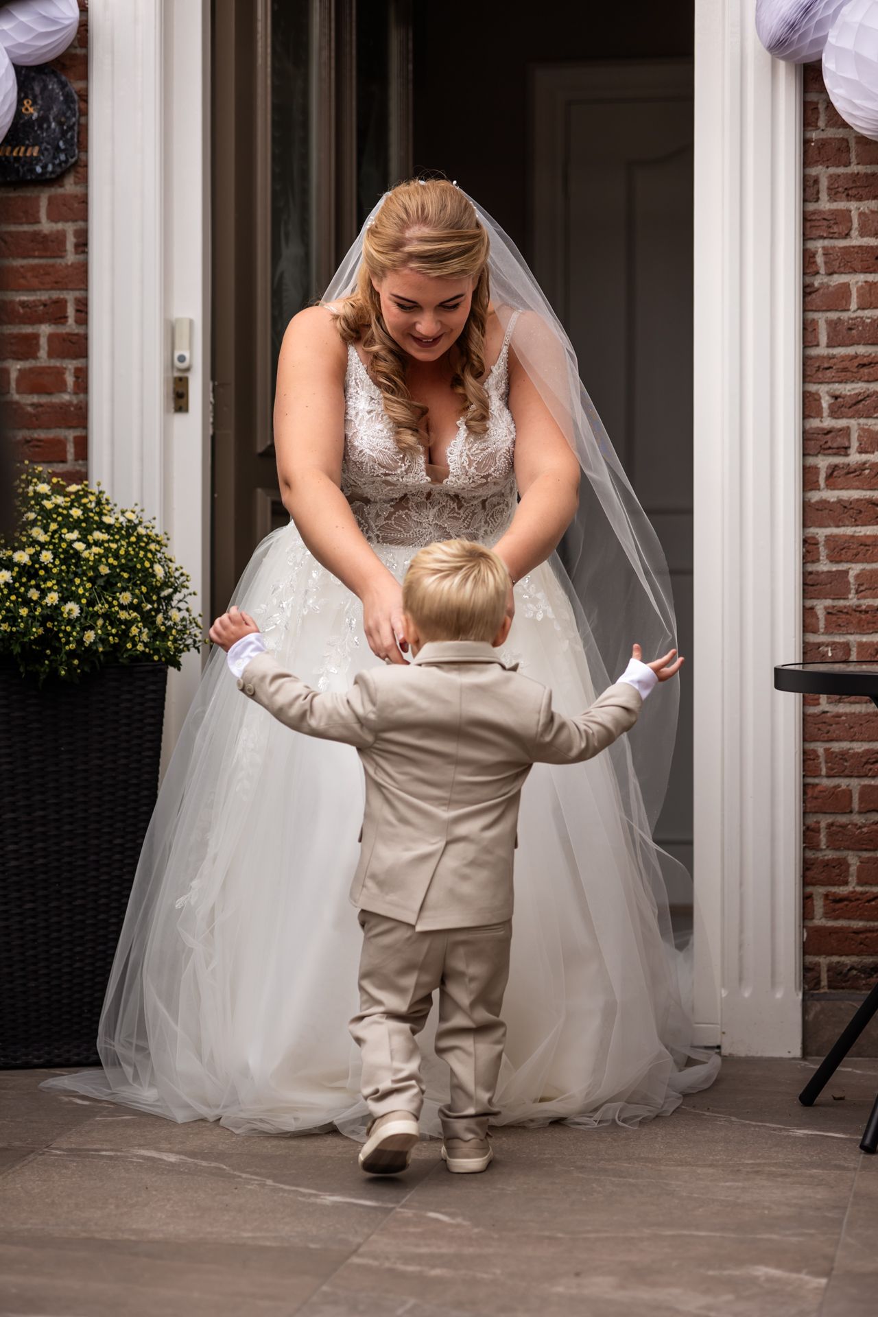 Bride in white gown bends down to interact with small child on porch.