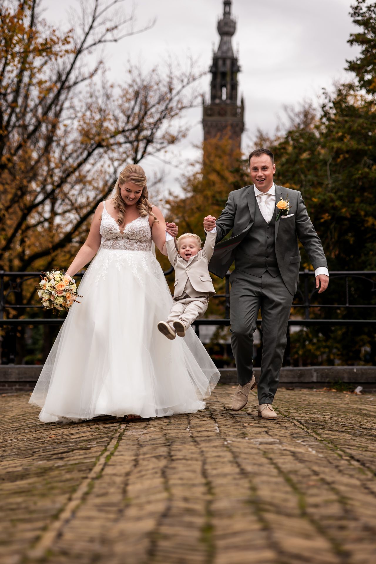 A wedding party celebrates on cobblestone path with historic church tower visible in autumn background.