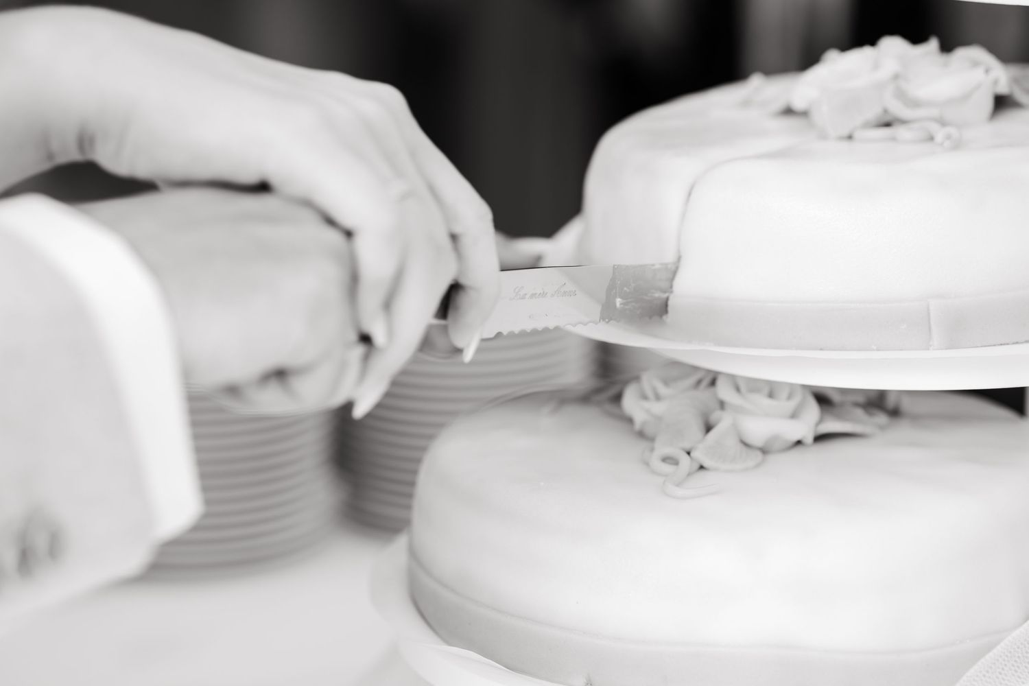 Black and white close-up of hands cutting into elegant white wedding cake tiers.