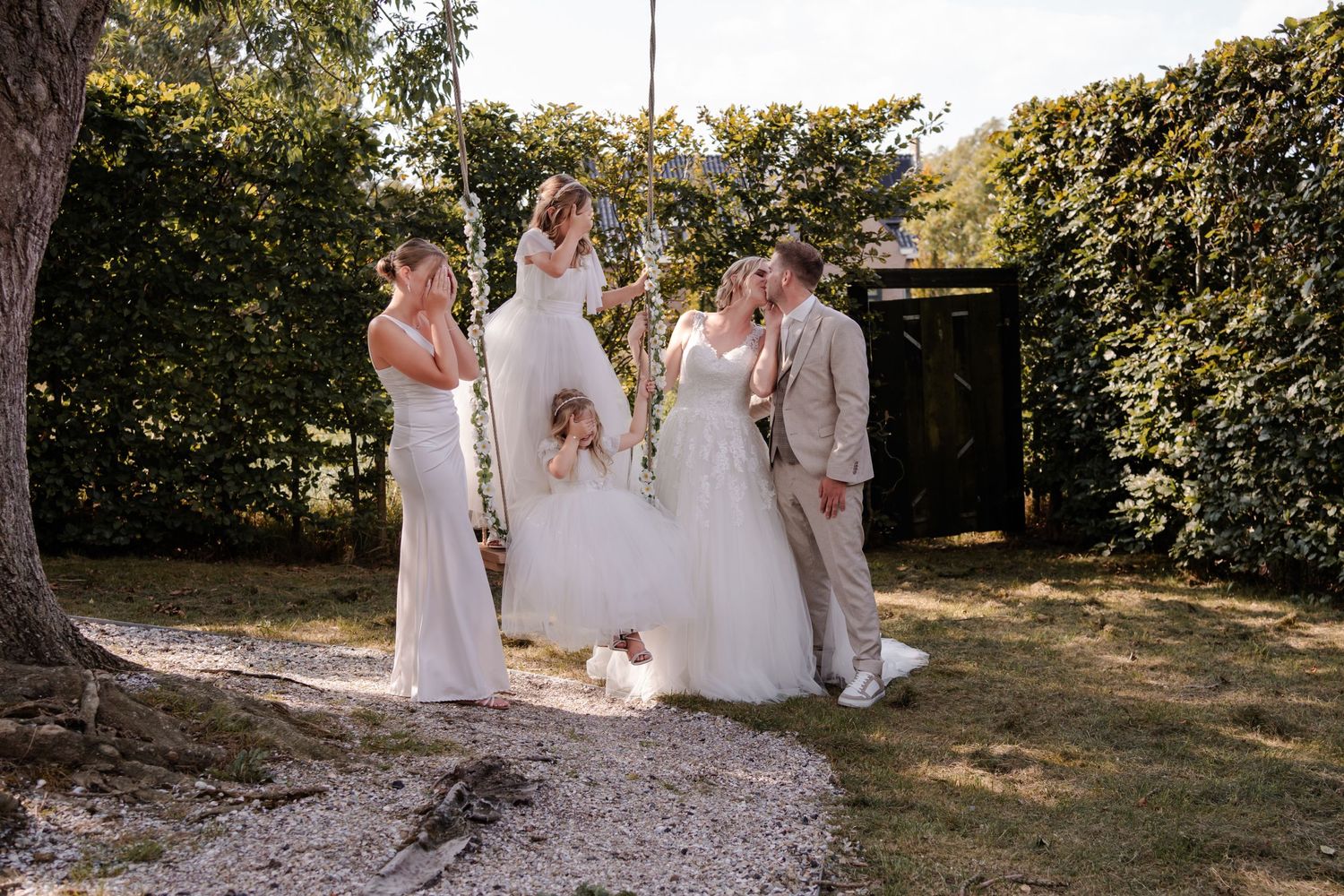 Wedding party gathered outdoors in a garden setting with sunlight filtering through the trees.