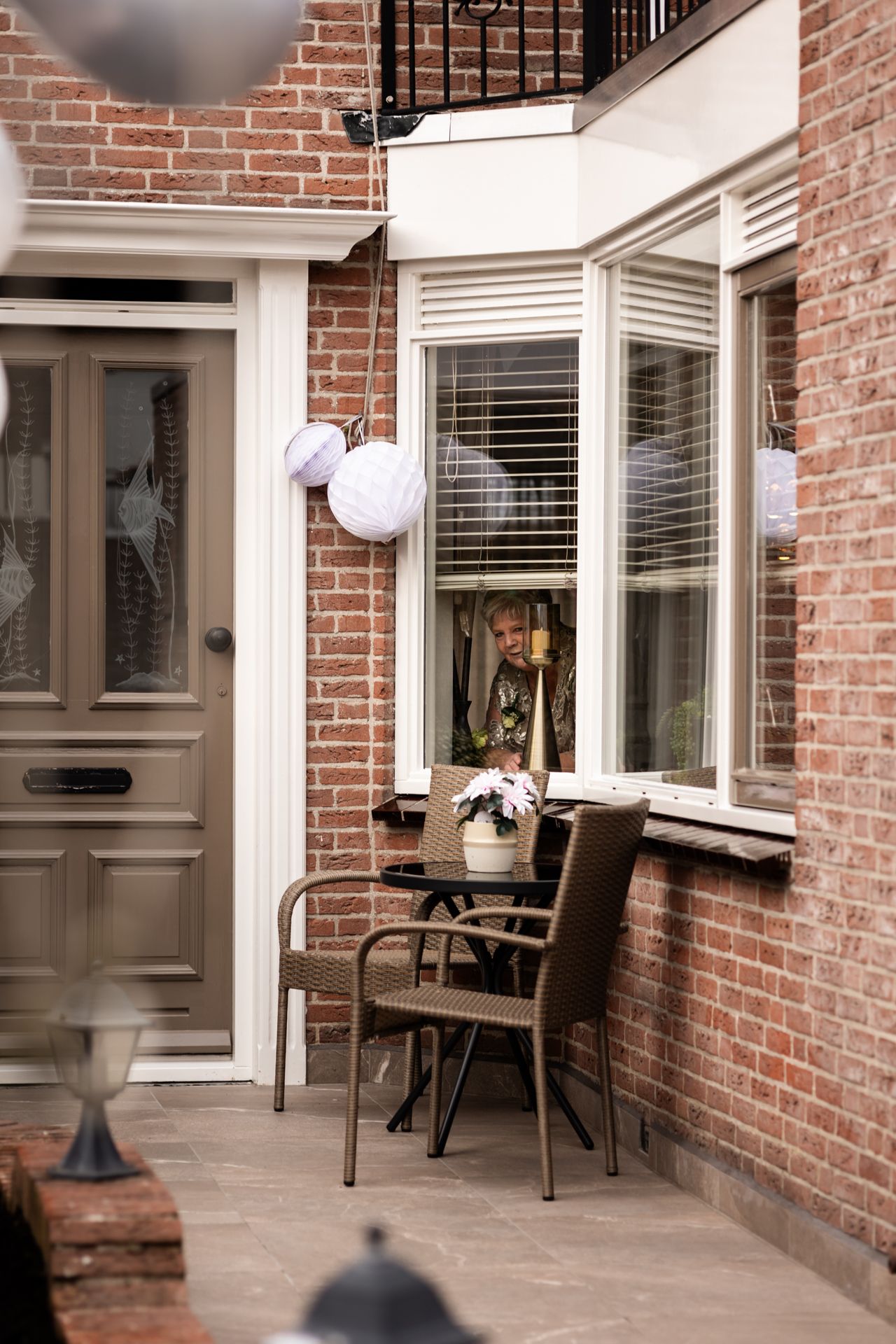 Brown brick house exterior with a porch chair and white paper lantern decorations framing a bay window.