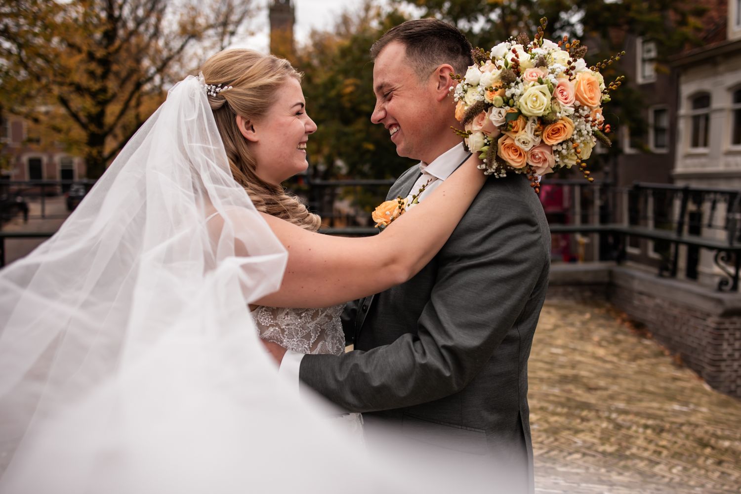 A bride and groom embrace while holding a peach and white floral bouquet on their wedding day.