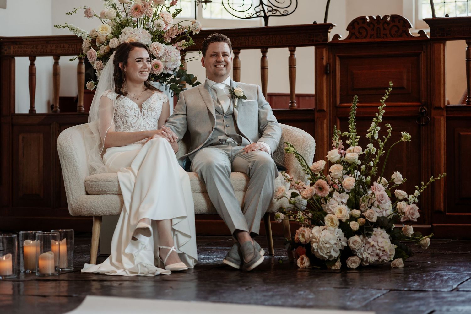 Bride and groom sitting together on a cream sofa surrounded by elegant floral arrangements during their wedding ceremony.