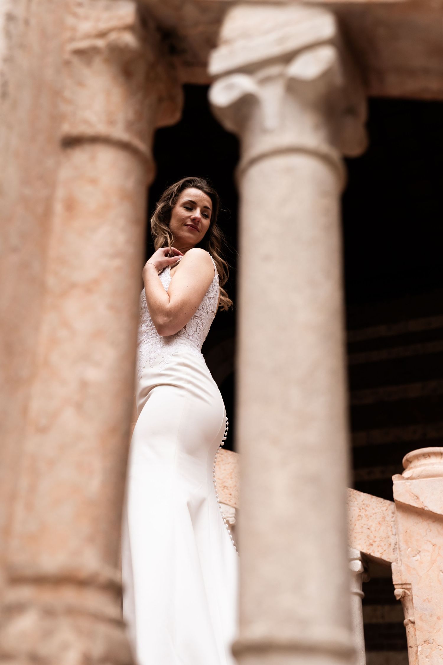 A bride in an elegant white dress poses between classic stone columns in a sepia-toned architectural setting.