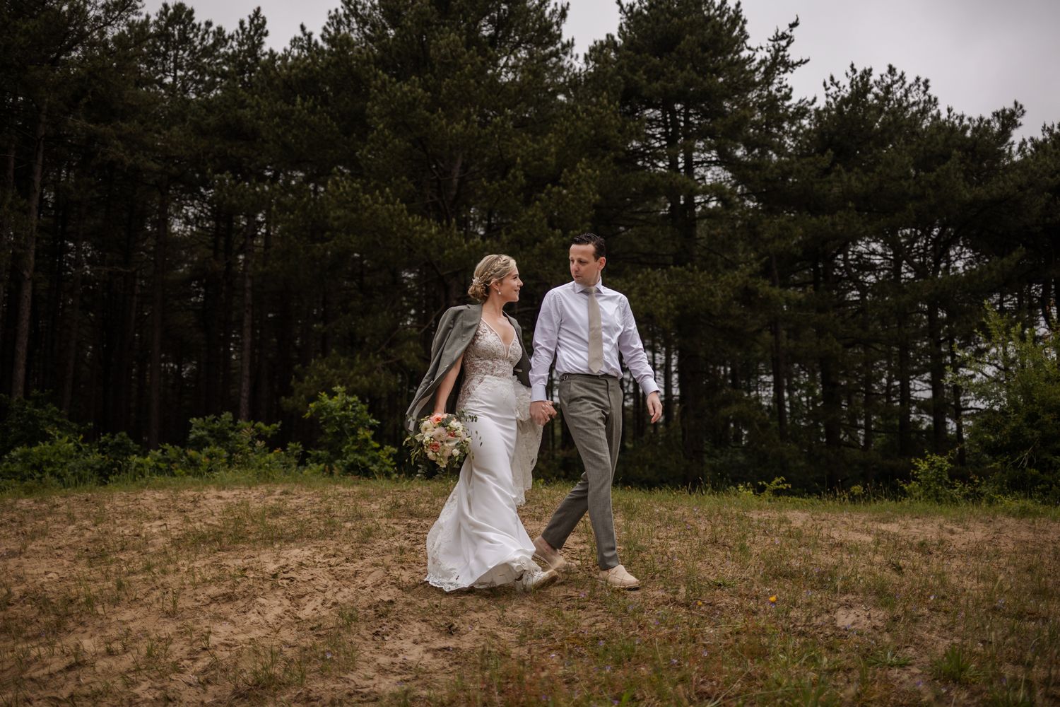A couple walks hand in hand through a clearing in a pine forest during their outdoor wedding photo session.