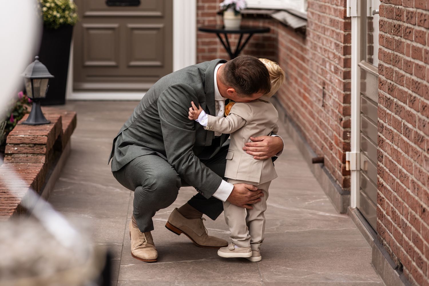 Parent kneeling to hug a small child wearing white on a brick pathway outside a house entrance.