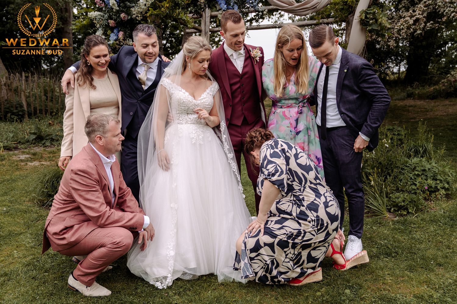 Wedding party poses outdoors on grass with bridesmaids in dusty rose dresses and floral patterns.