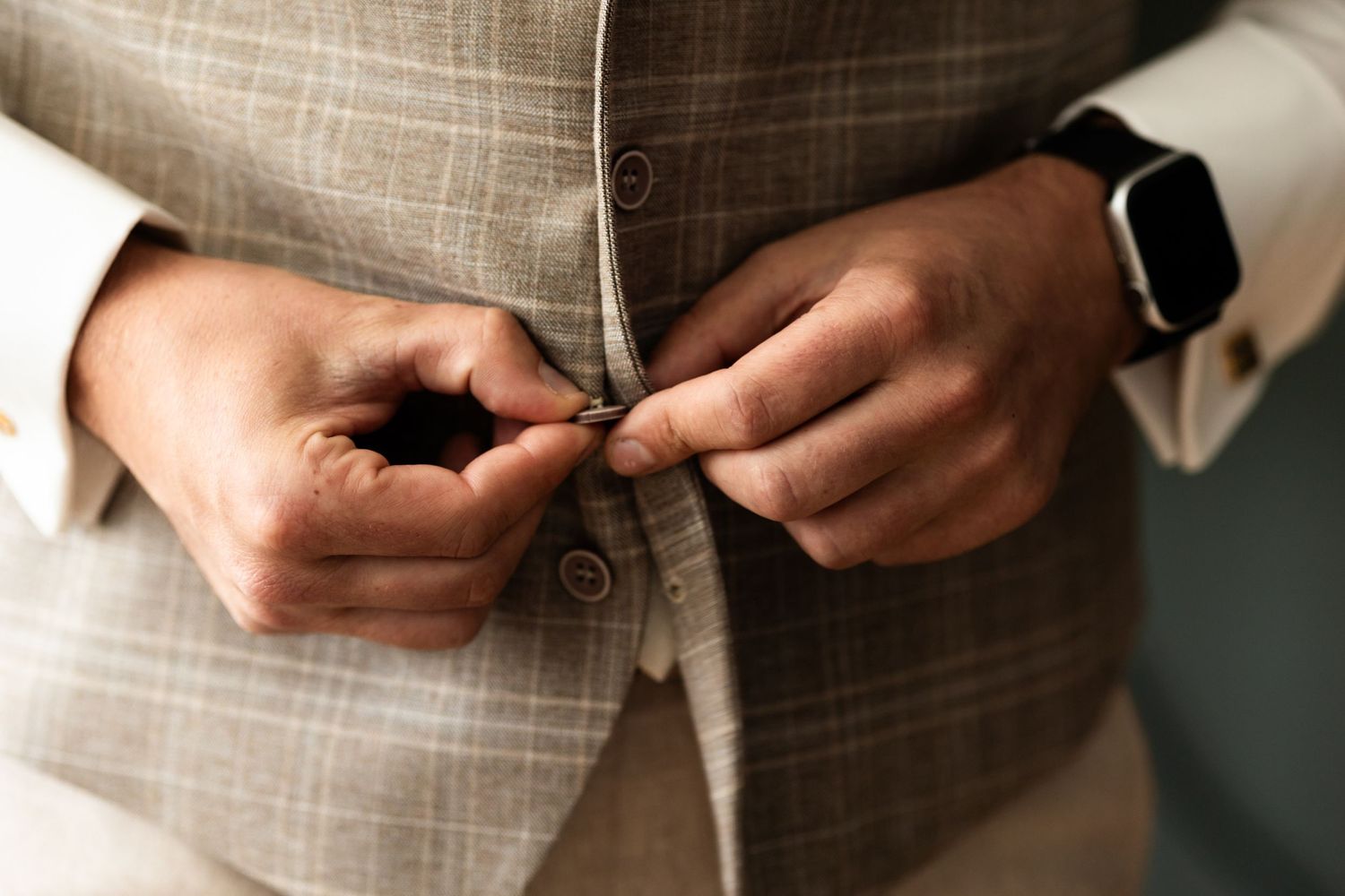 Close-up view of hands buttoning a beige linen suit vest with a smartwatch visible on the wrist.