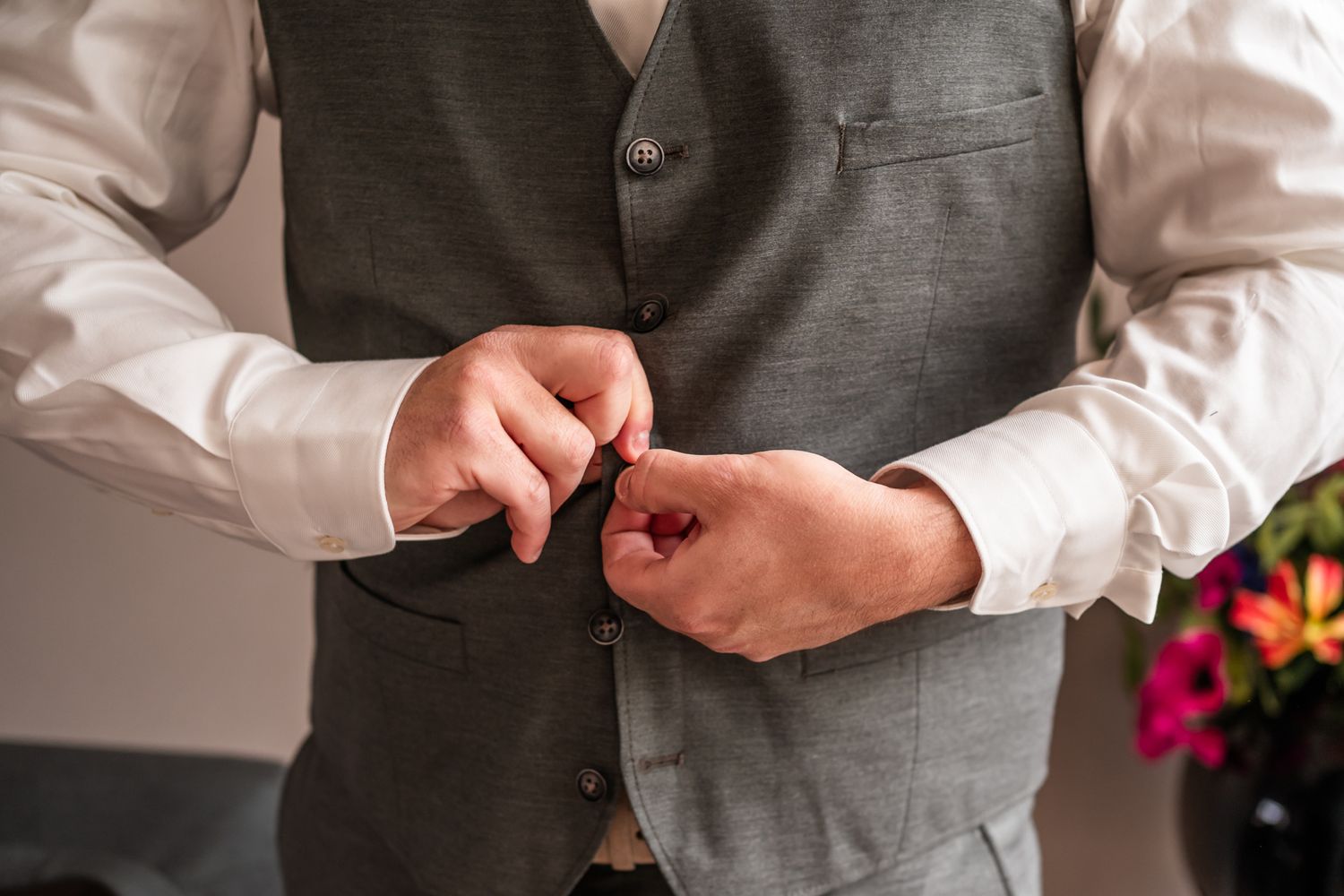 Groom adjusts his gray vest buttons while preparing for wedding ceremony.