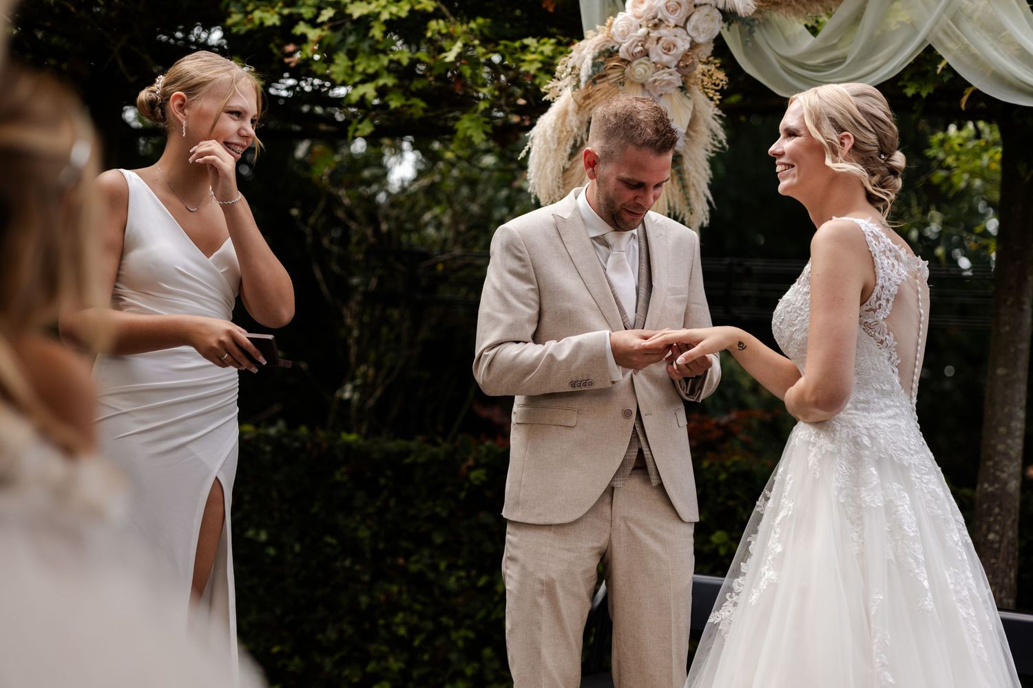 An intimate outdoor wedding ceremony with the couple exchanging rings under green trees.