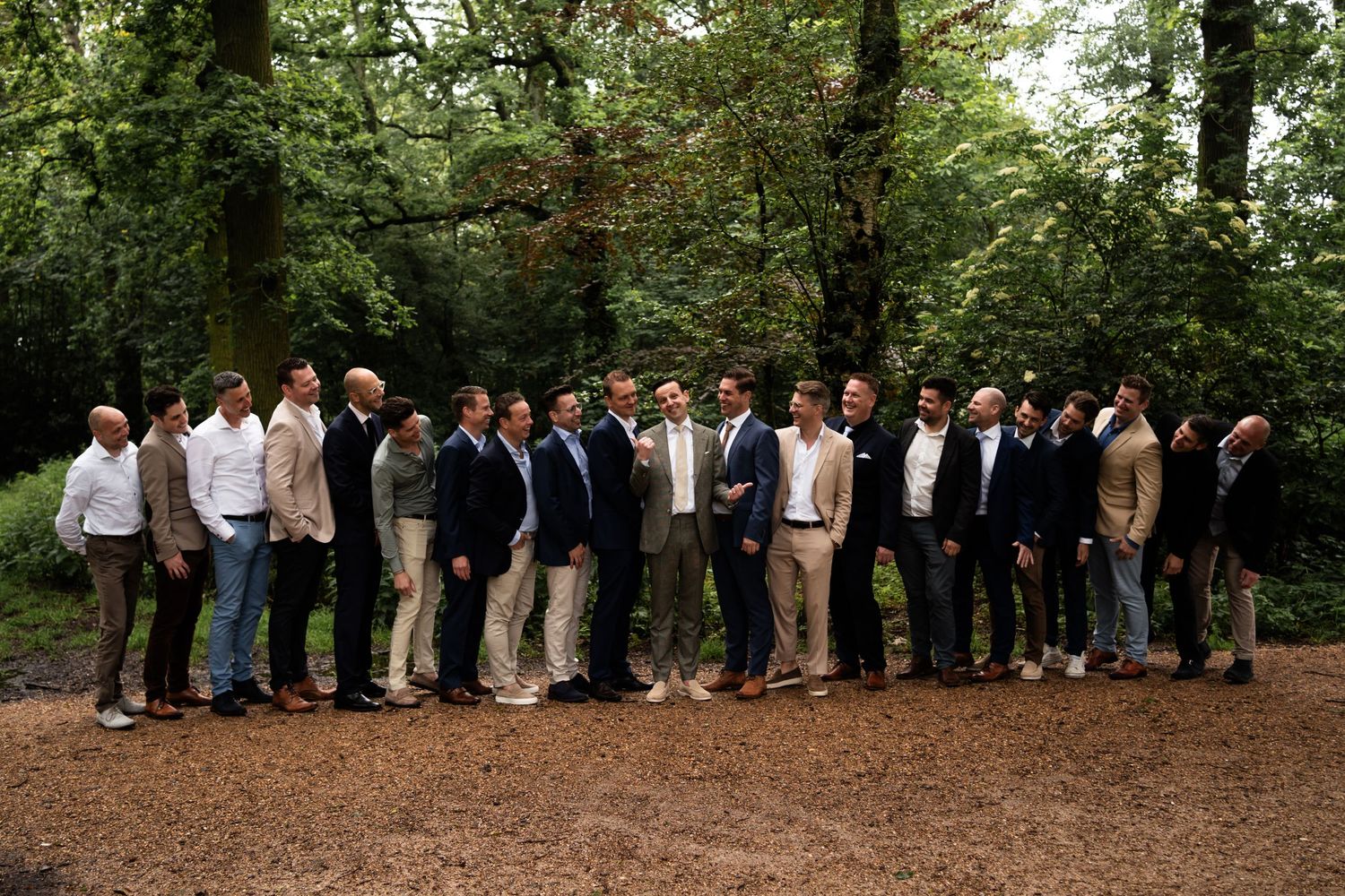 A large group of groomsmen in casual formal attire pose together in a wooded setting.