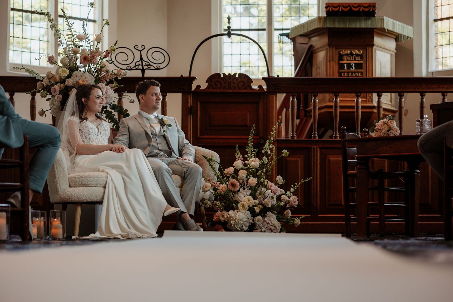 Bride and groom seated together during wedding ceremony in elegant wood-paneled chapel with floral arrangements.