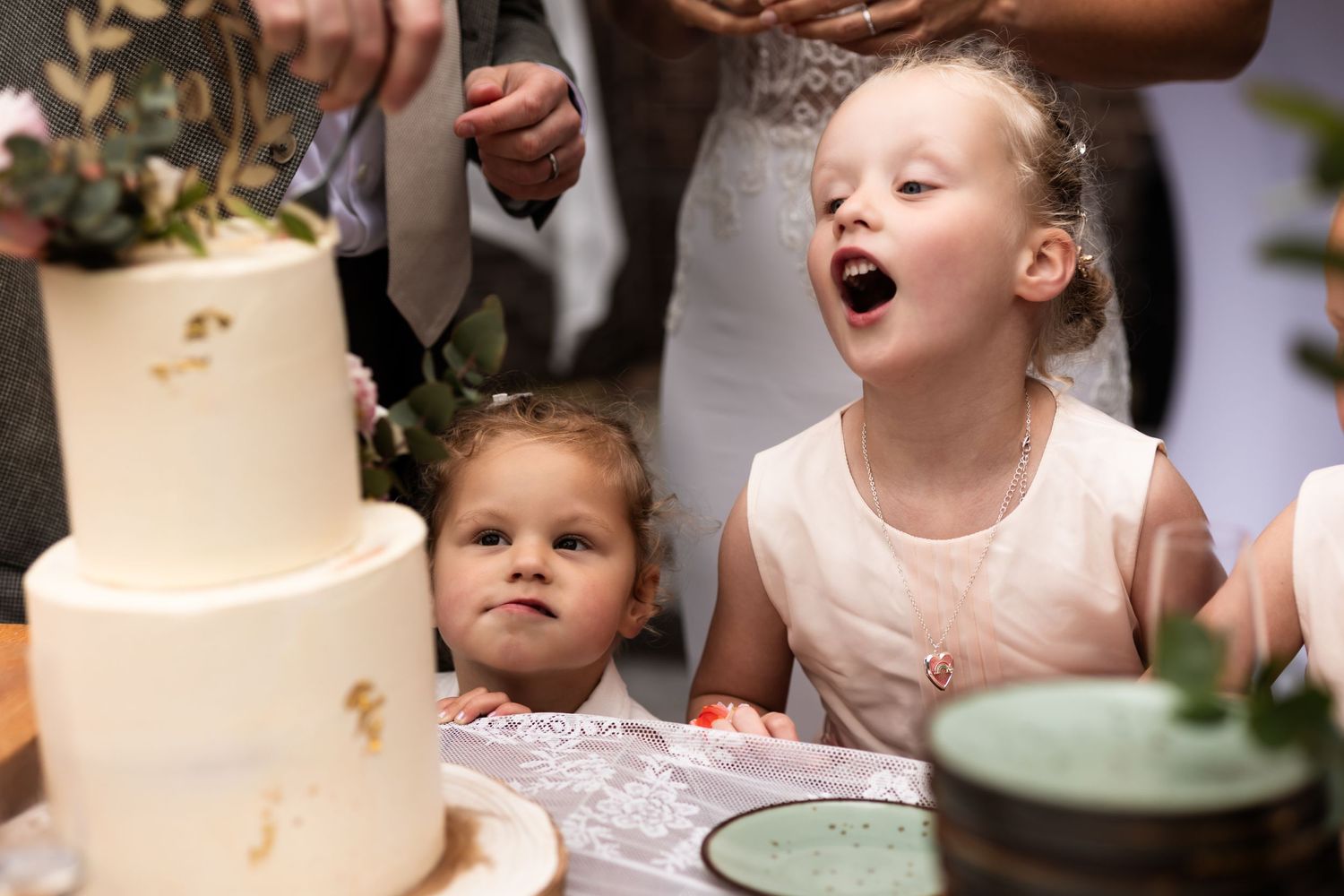 Two excited young children eagerly watch a white wedding cake being cut.