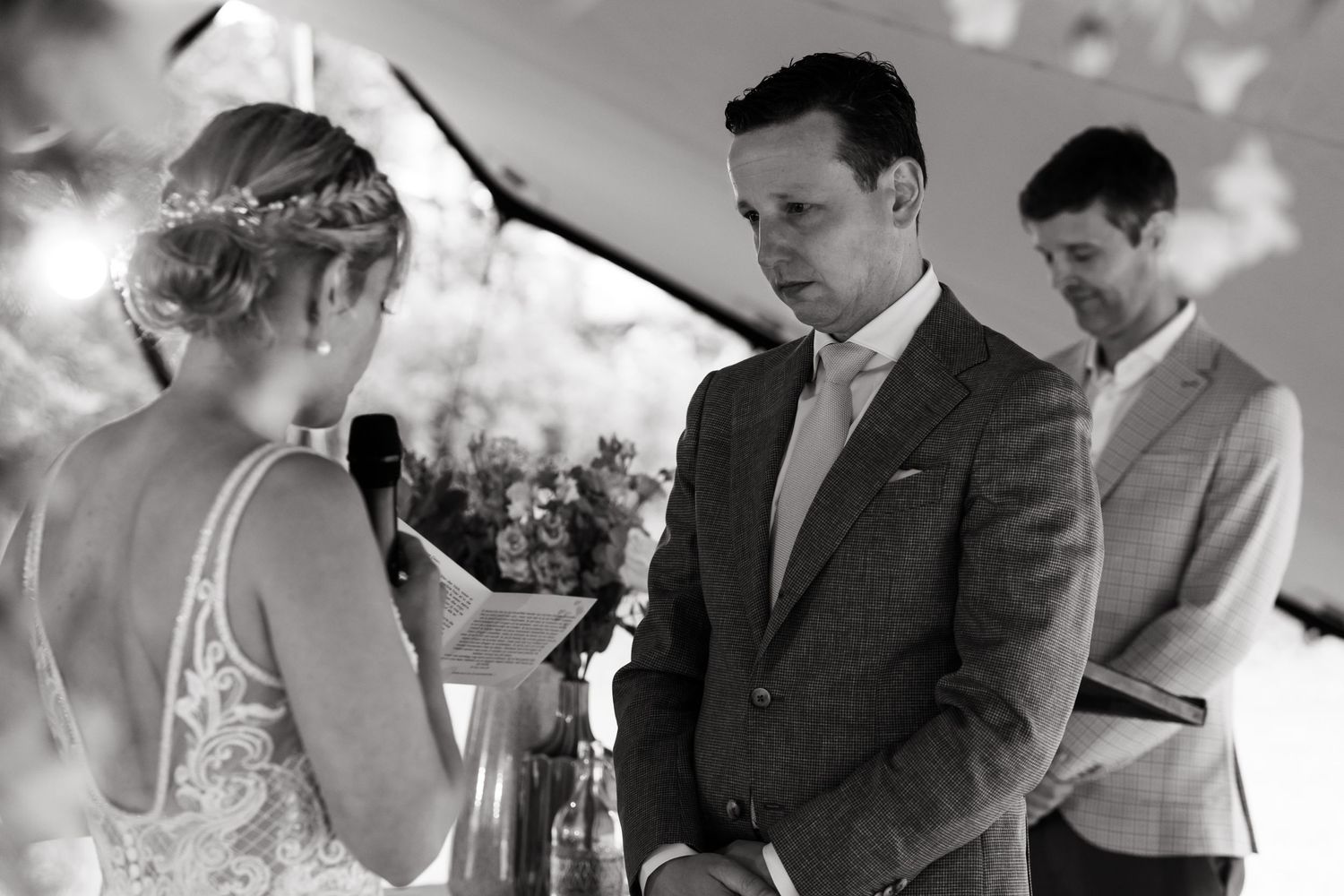 Black and white photo of wedding ceremony participants exchanging vows under outdoor tent lighting.