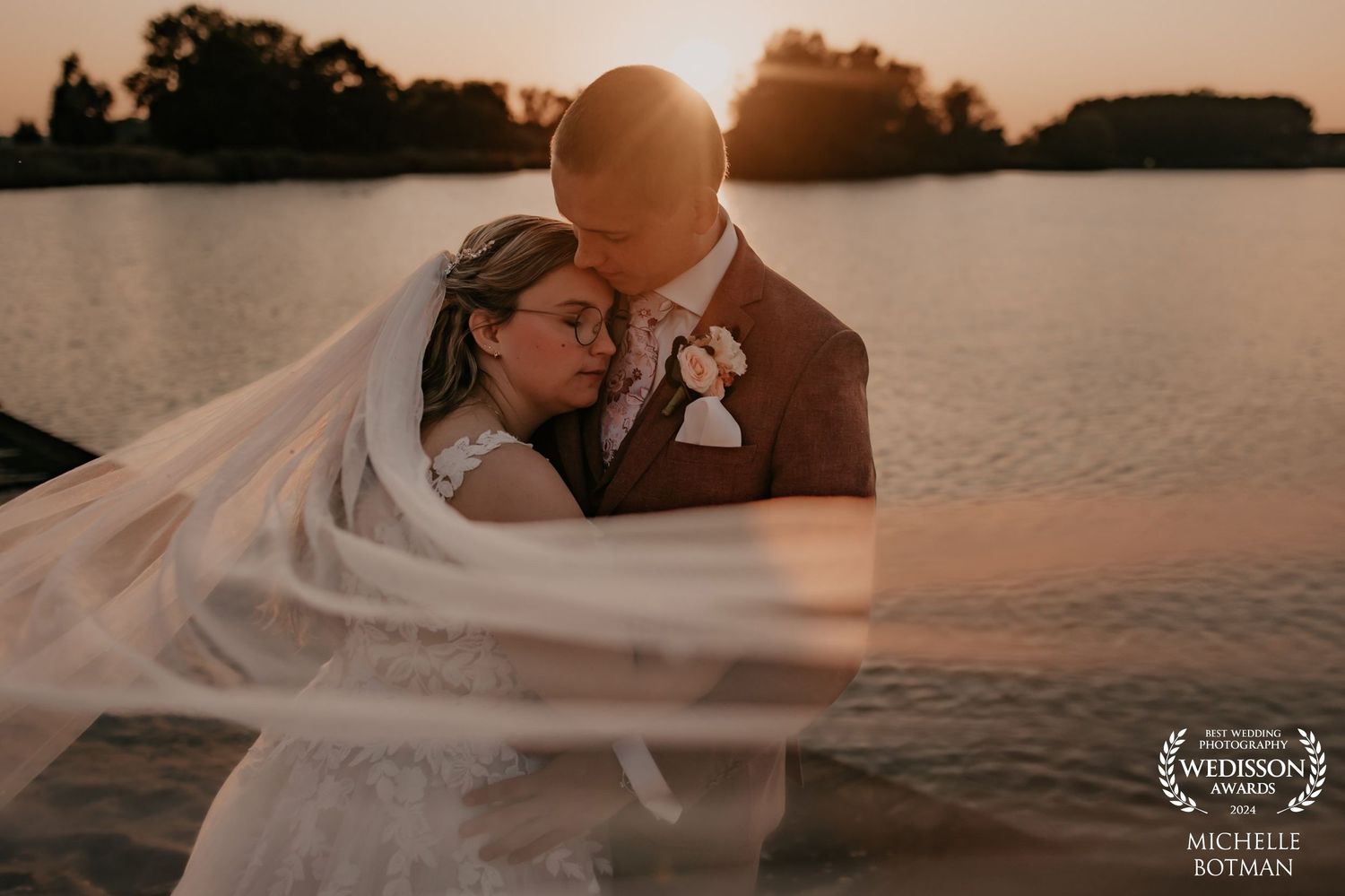 Romantic sunset wedding portrait by lake with flowing veil in gentle breeze.