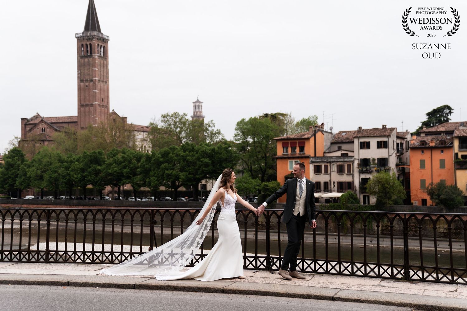 A bride and groom pose on an ornate bridge in Verona, Italy with a historic church spire and colorful buildings in background.
