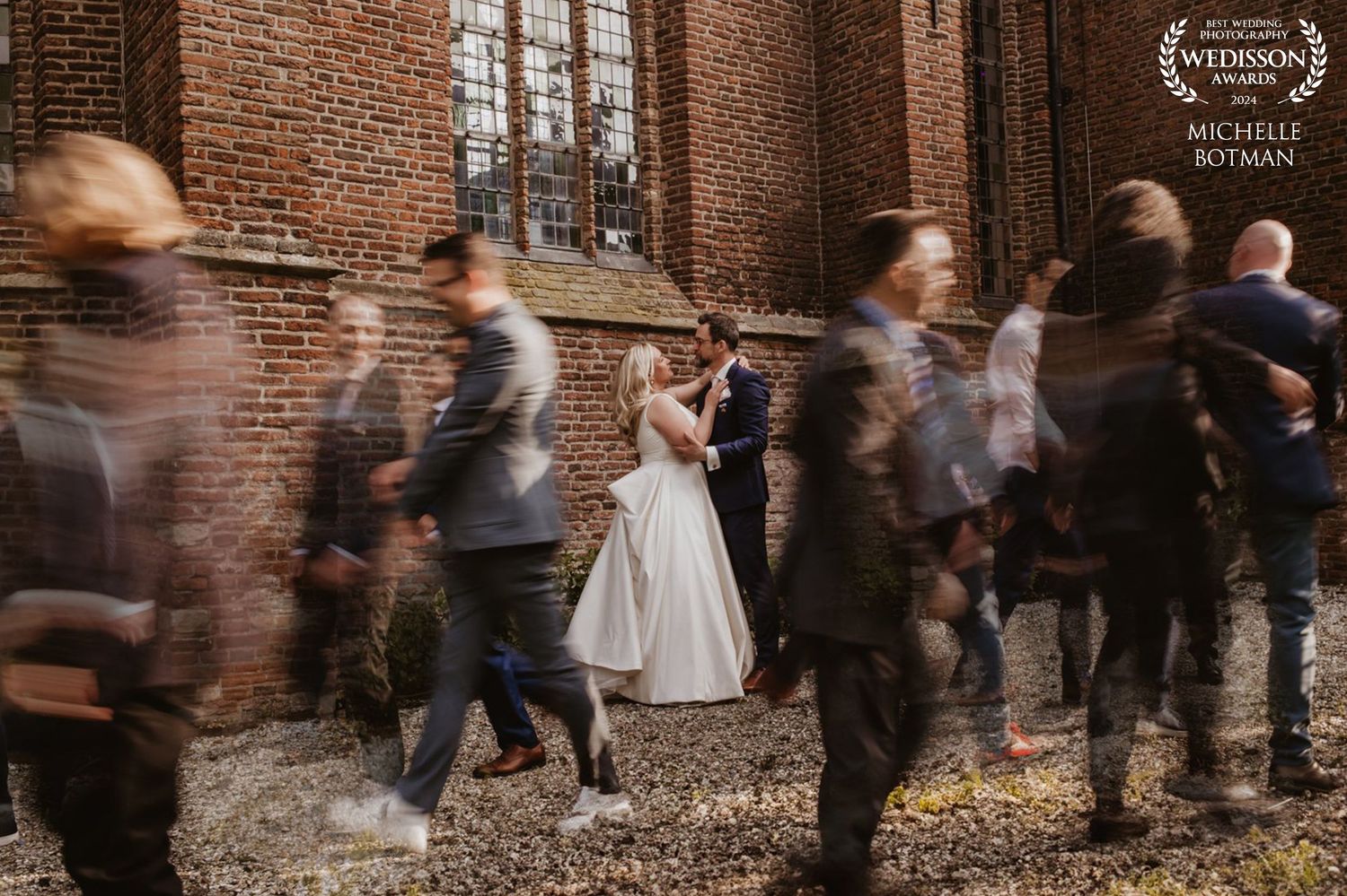 Motion blur of wedding guests moving past couple in brick courtyard.