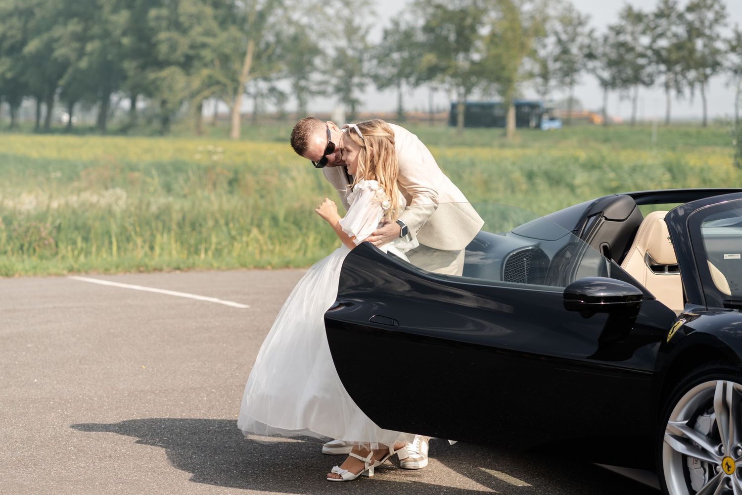 A couple leans over a black luxury convertible car parked in a countryside setting with wildflowers in the background.