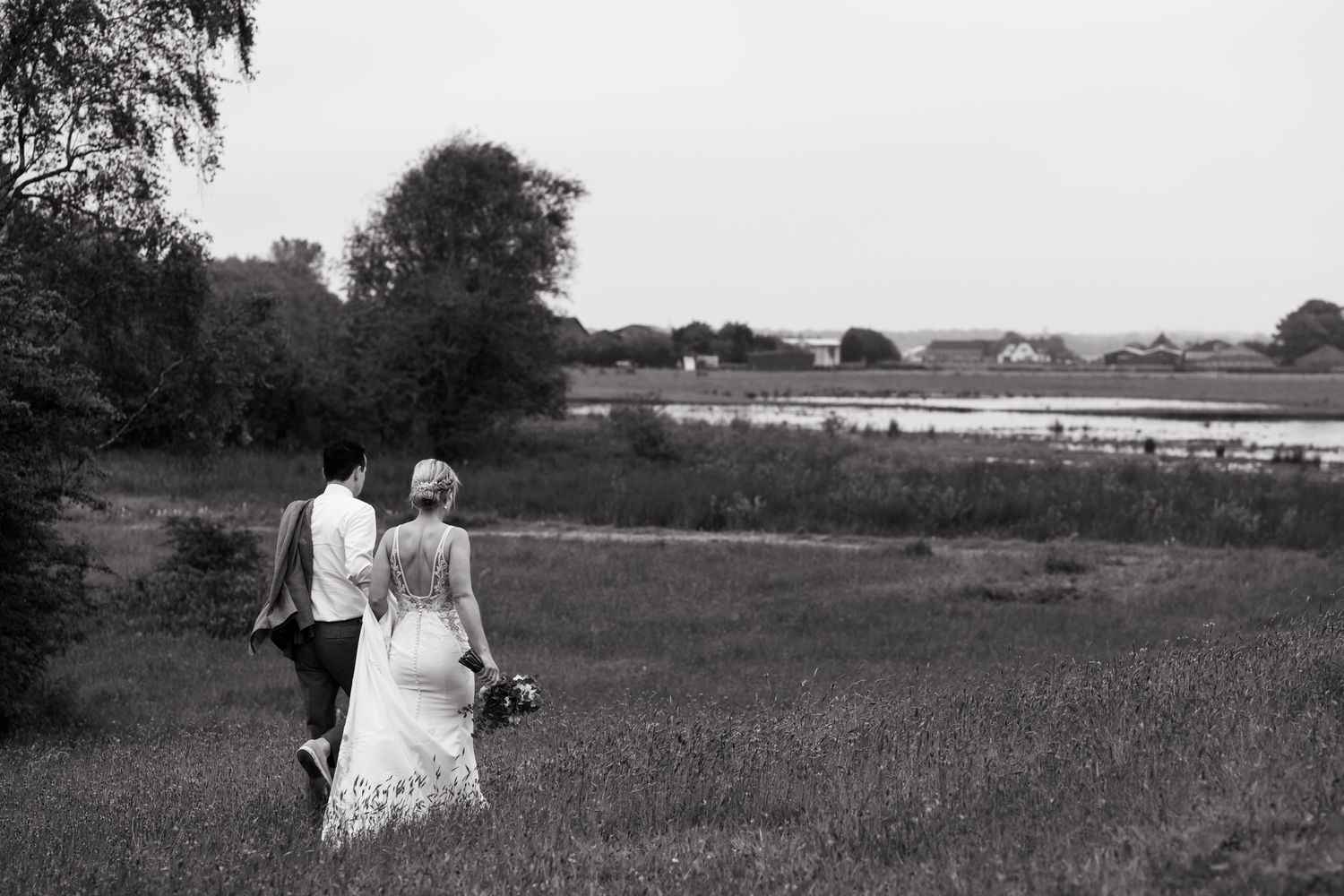 A couple walks through a meadow with a scenic lake view in black and white photography.