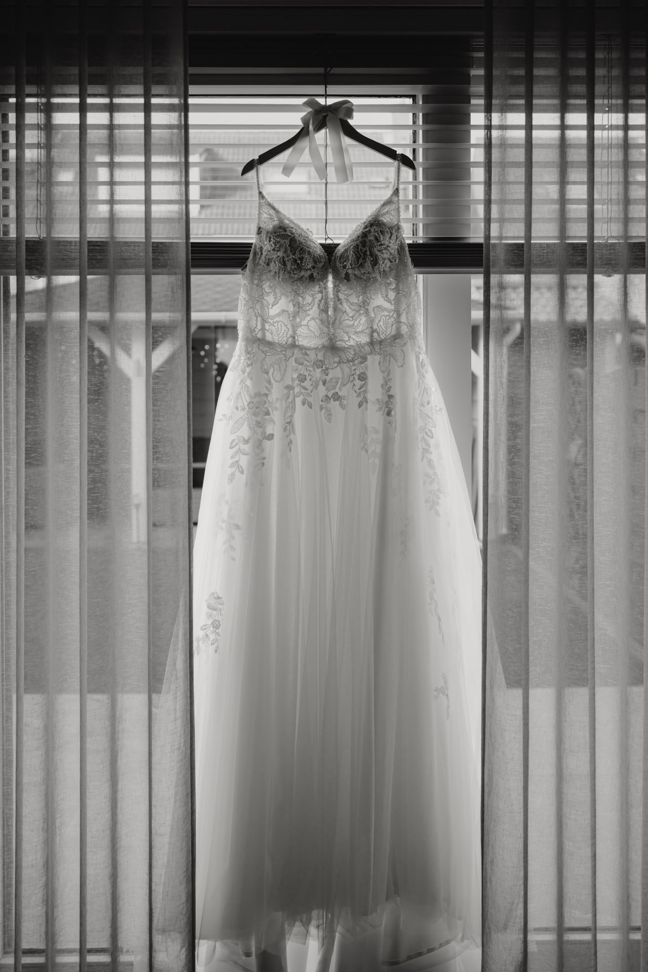 An elegant white lace wedding dress with a sweetheart neckline hangs on a wooden hanger against sheer curtains.