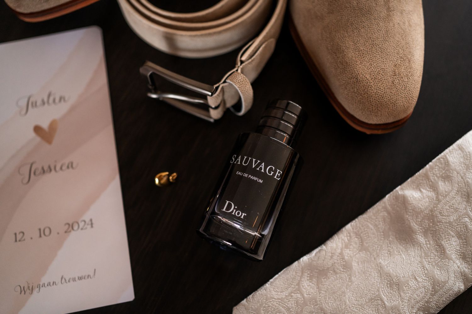 Wedding invitation, brown leather belt, Dior Homme cologne bottle and suede shoe arranged on black surface with lace detail.