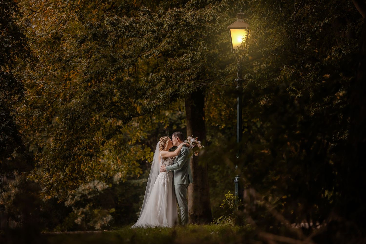A couple in wedding attire shares a romantic moment under glowing lanterns in a lush garden setting at dusk.