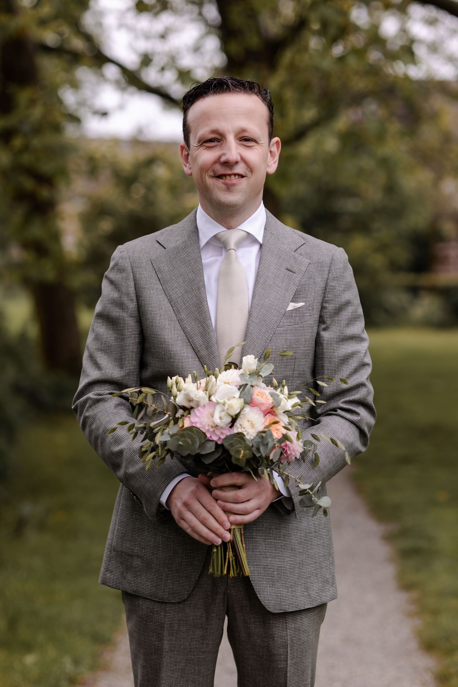 A groom in a grey suit holds a delicate bouquet of pink and white flowers while standing on a garden path.