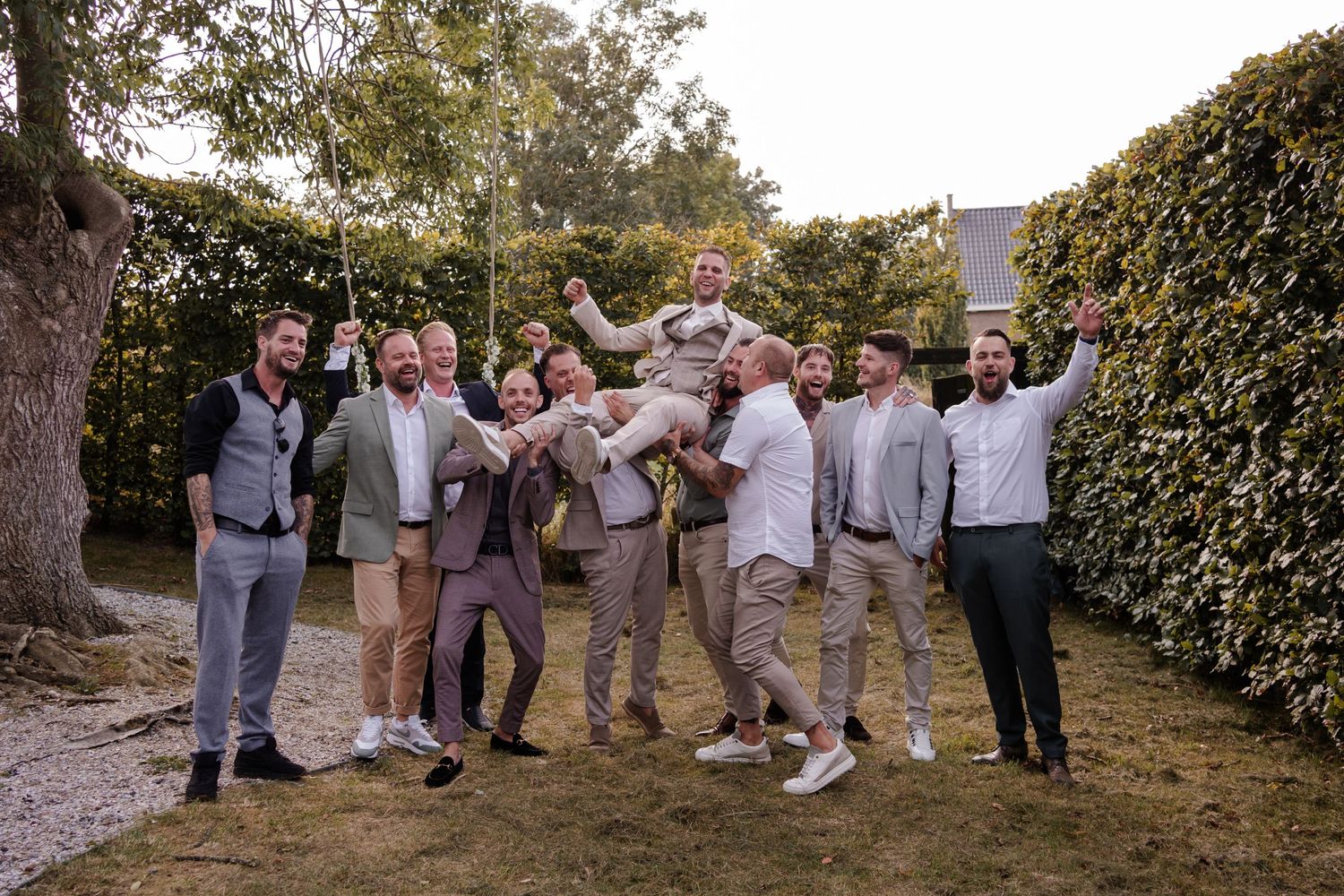 Groomsmen in casual gray and beige attire celebrate joyfully outdoors during a garden wedding photo session.