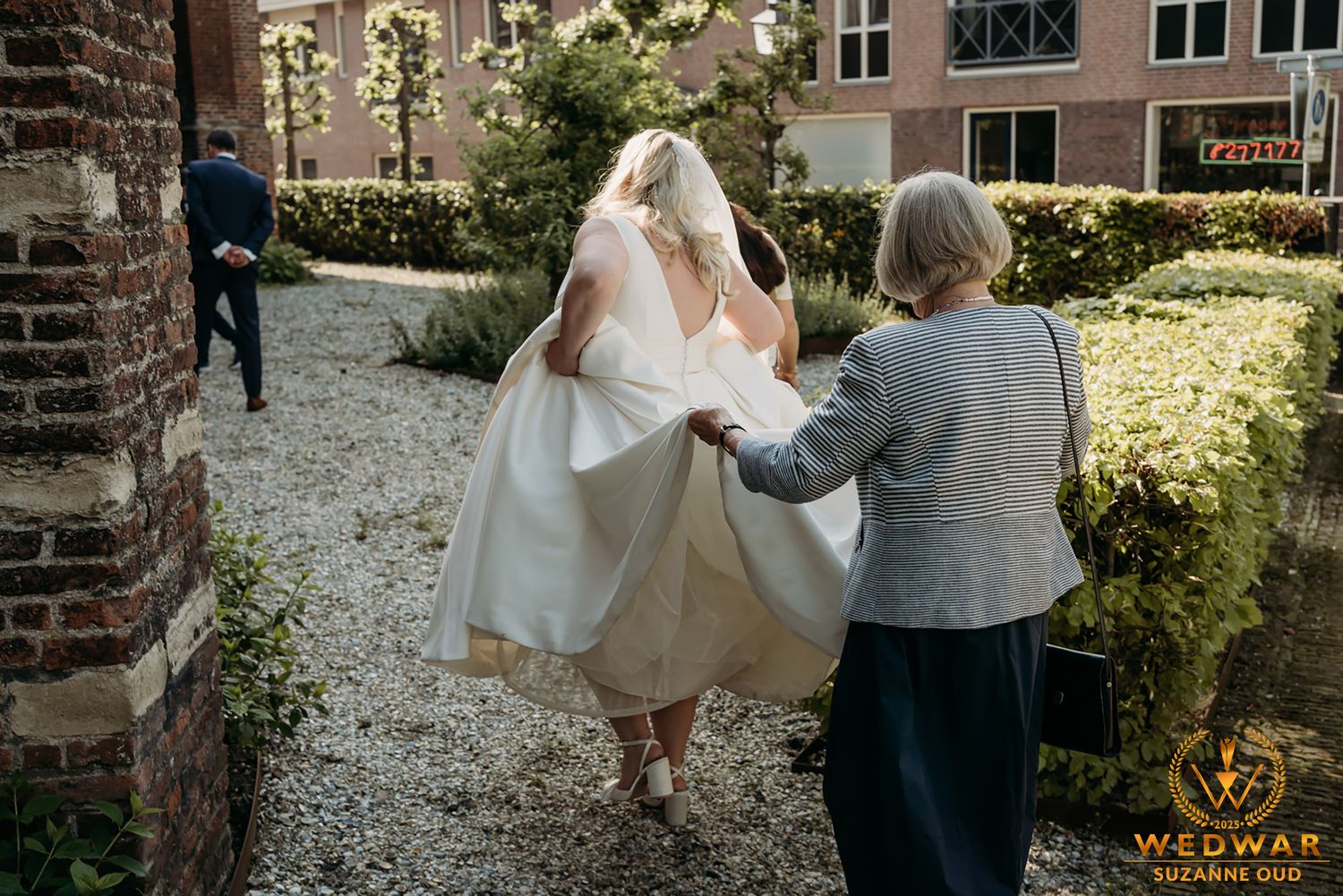 Wedding party members walk together through a garden setting with brick walls and greenery in the background.