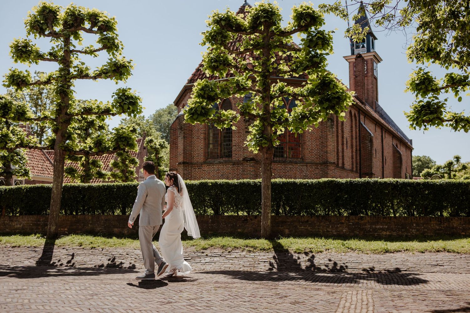 Bride and groom walking hand in hand past trimmed trees and a brick church on a sunny wedding day.