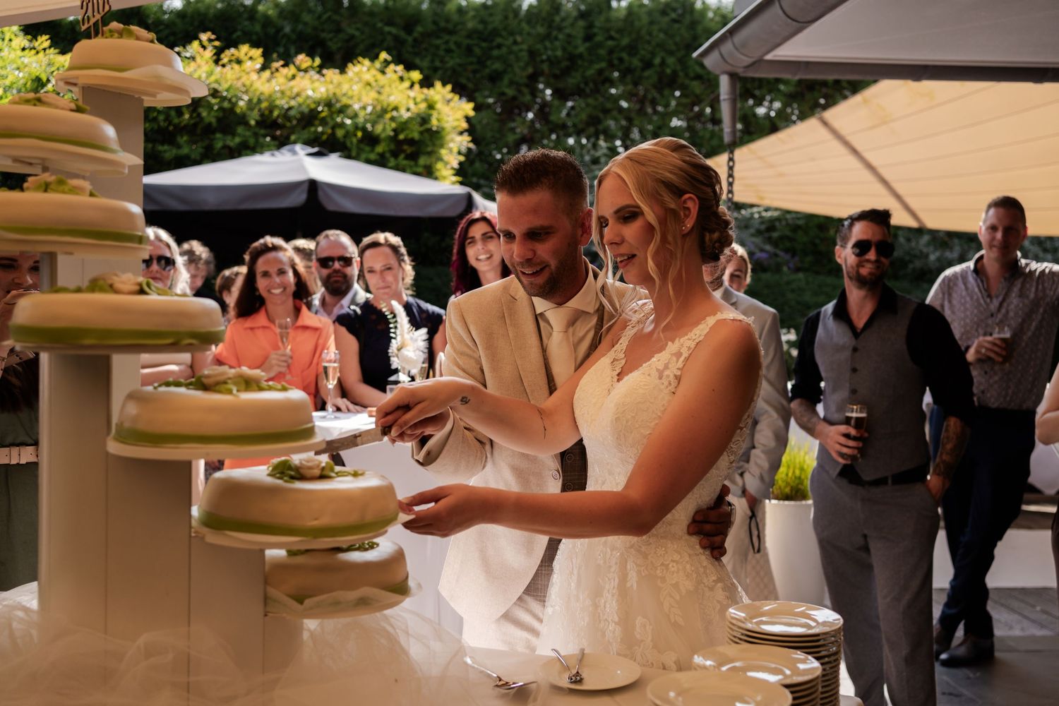 A couple cutting their multi-tiered wedding cake together at an outdoor reception while guests watch in the background.