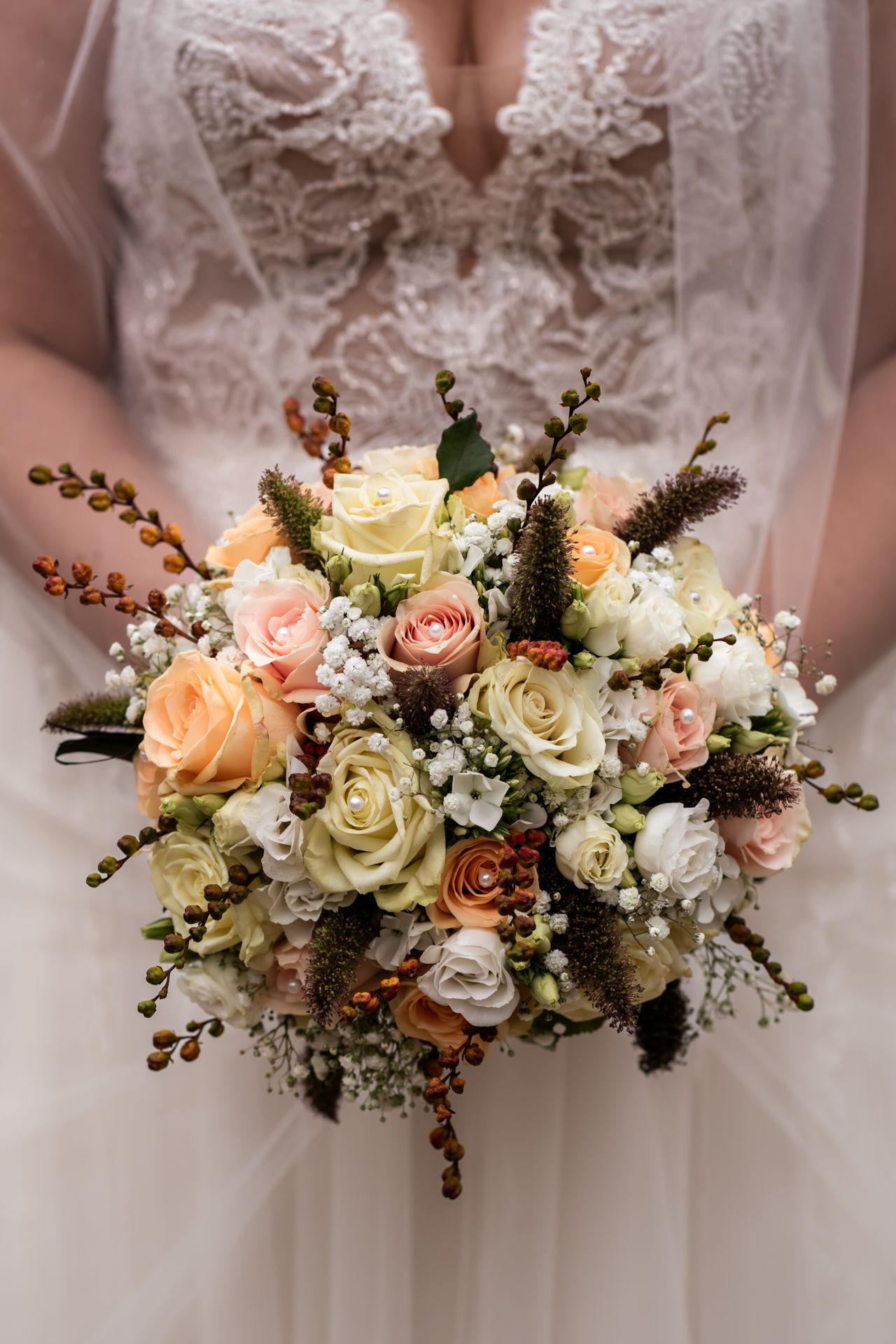 Elegant wedding bouquet with peach and cream roses, white flowers, berries and greenery against lace dress detail.