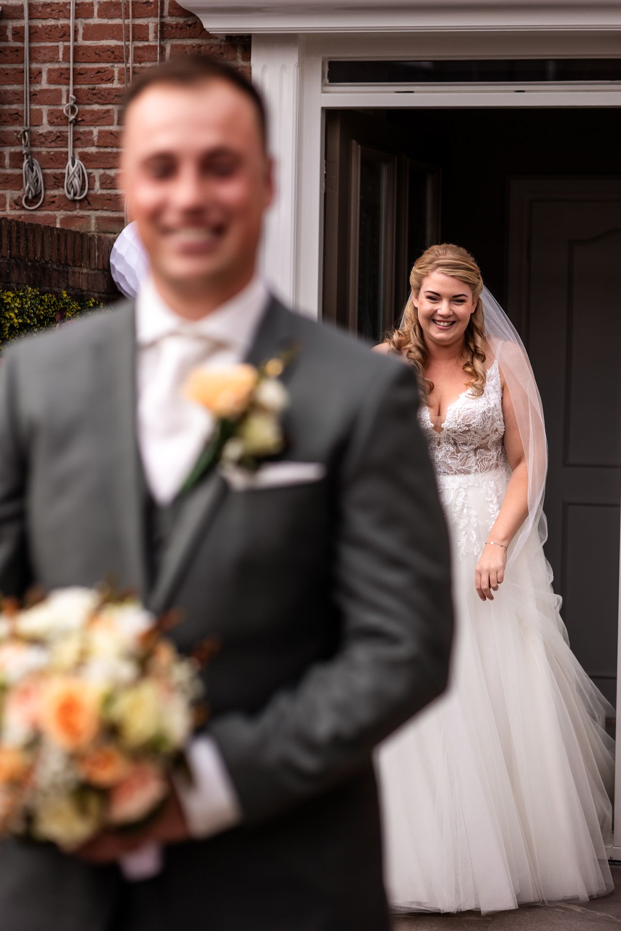 A wedding first look moment with bride in elegant white dress standing behind groom in grey suit with boutonniere.