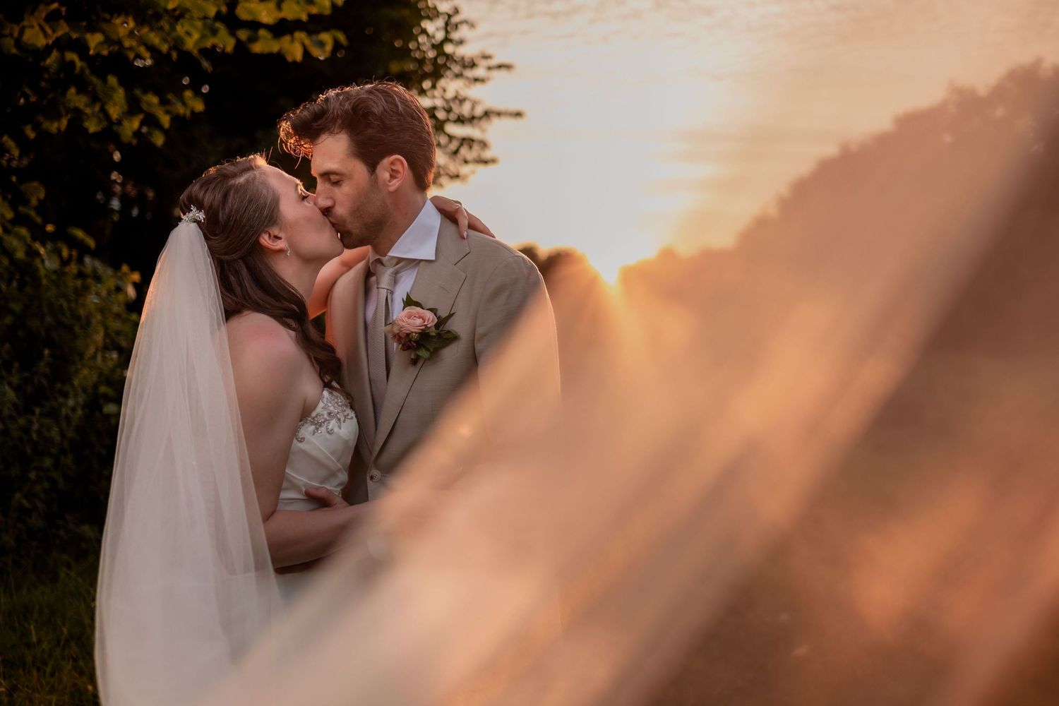 A romantic wedding couple shares a tender moment at sunset, with the bride's veil flowing in the foreground.