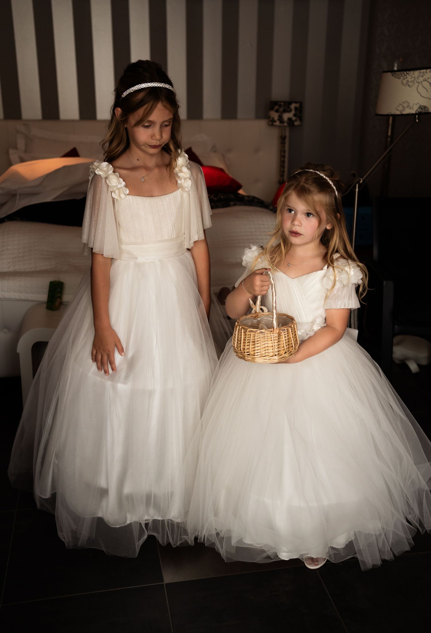 Two young flower girls in flowing white tulle dresses stand together, one holding a small basket.