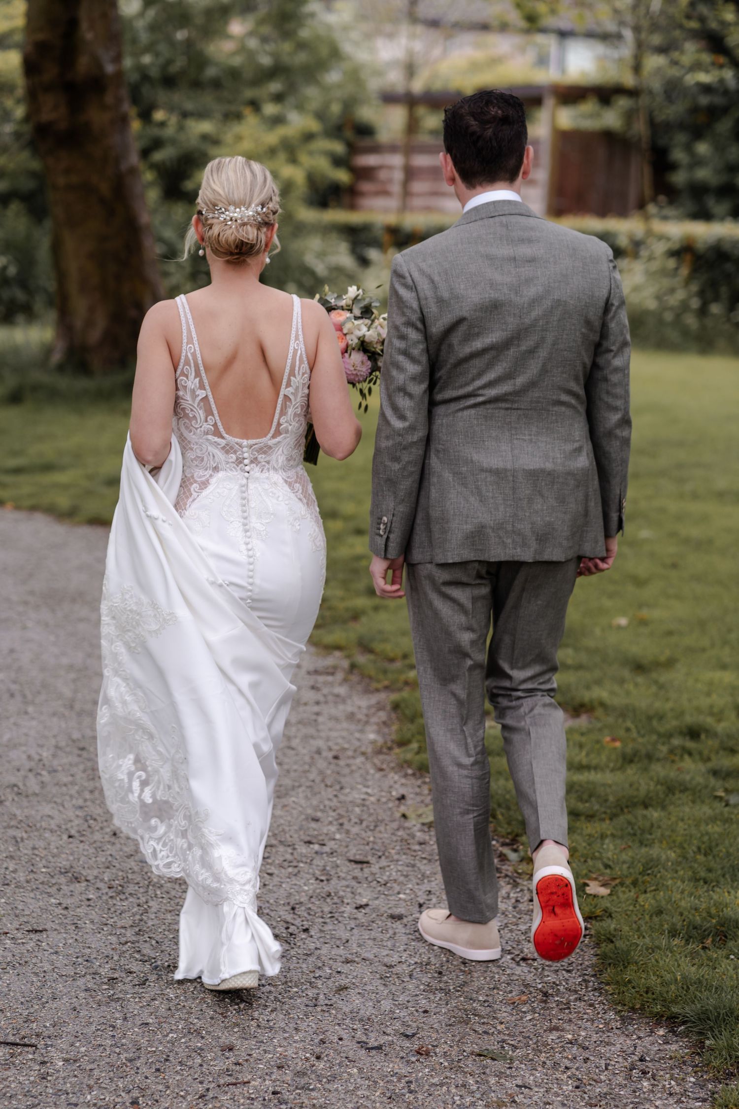 A wedding couple walks down a garden path, with the bride in a lace backless gown and groom in a gray suit.