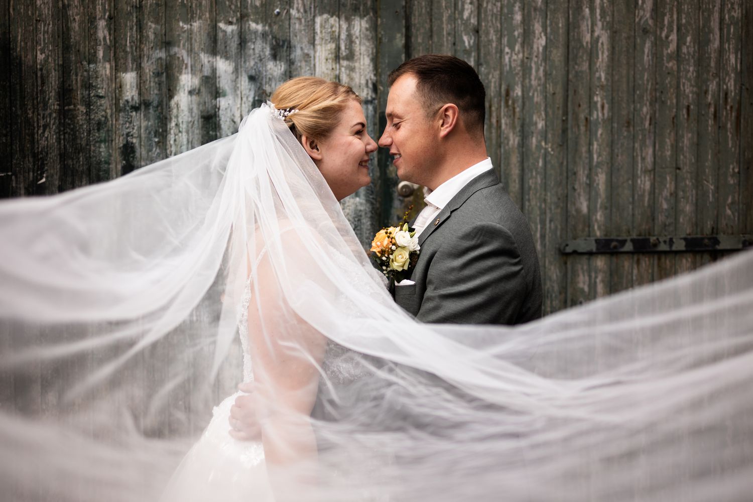 A dreamy wedding portrait with a flowing veil creating movement against a rustic wooden backdrop.