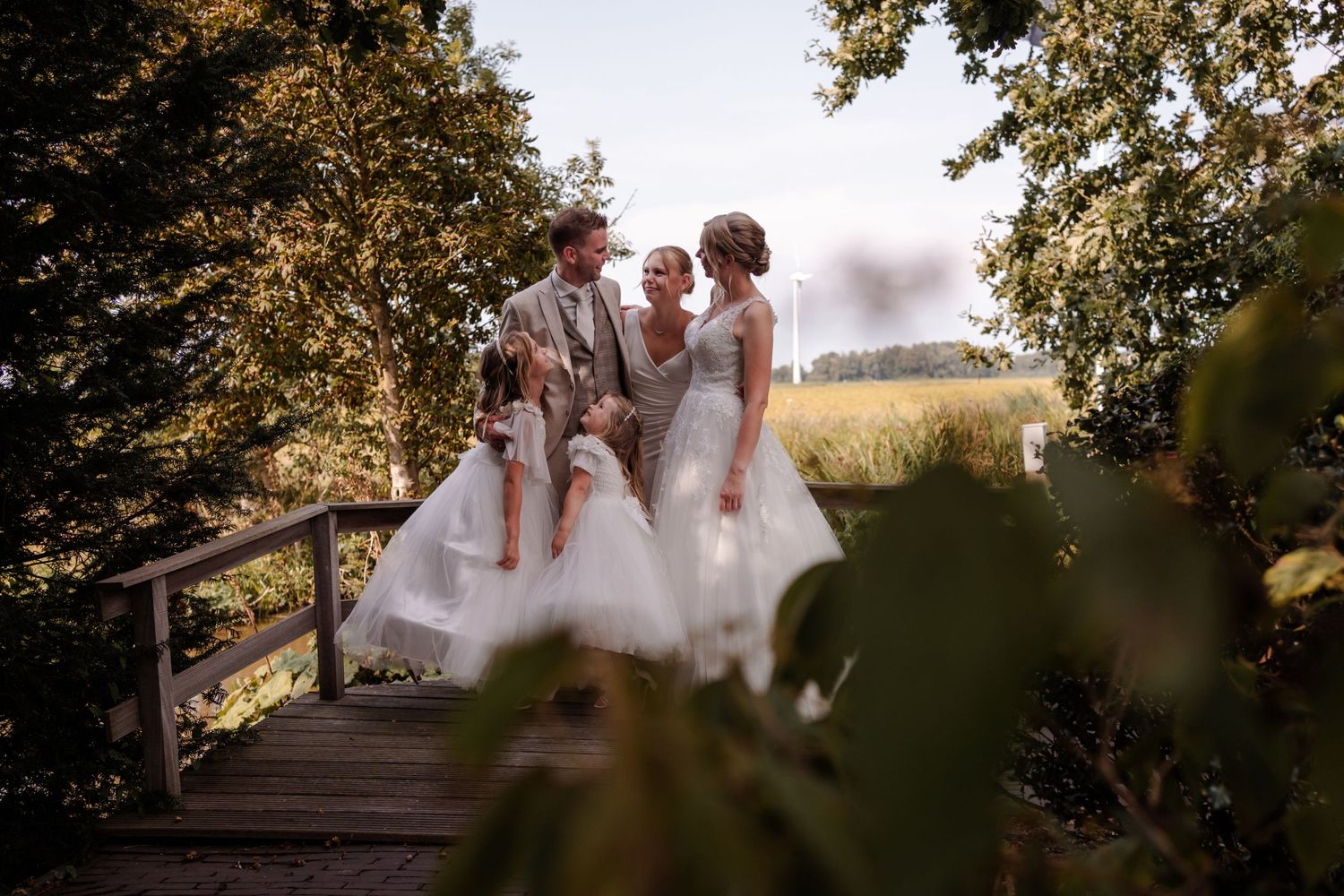 Three people in white dresses walking across a wooden bridge surrounded by trees on a sunny afternoon.