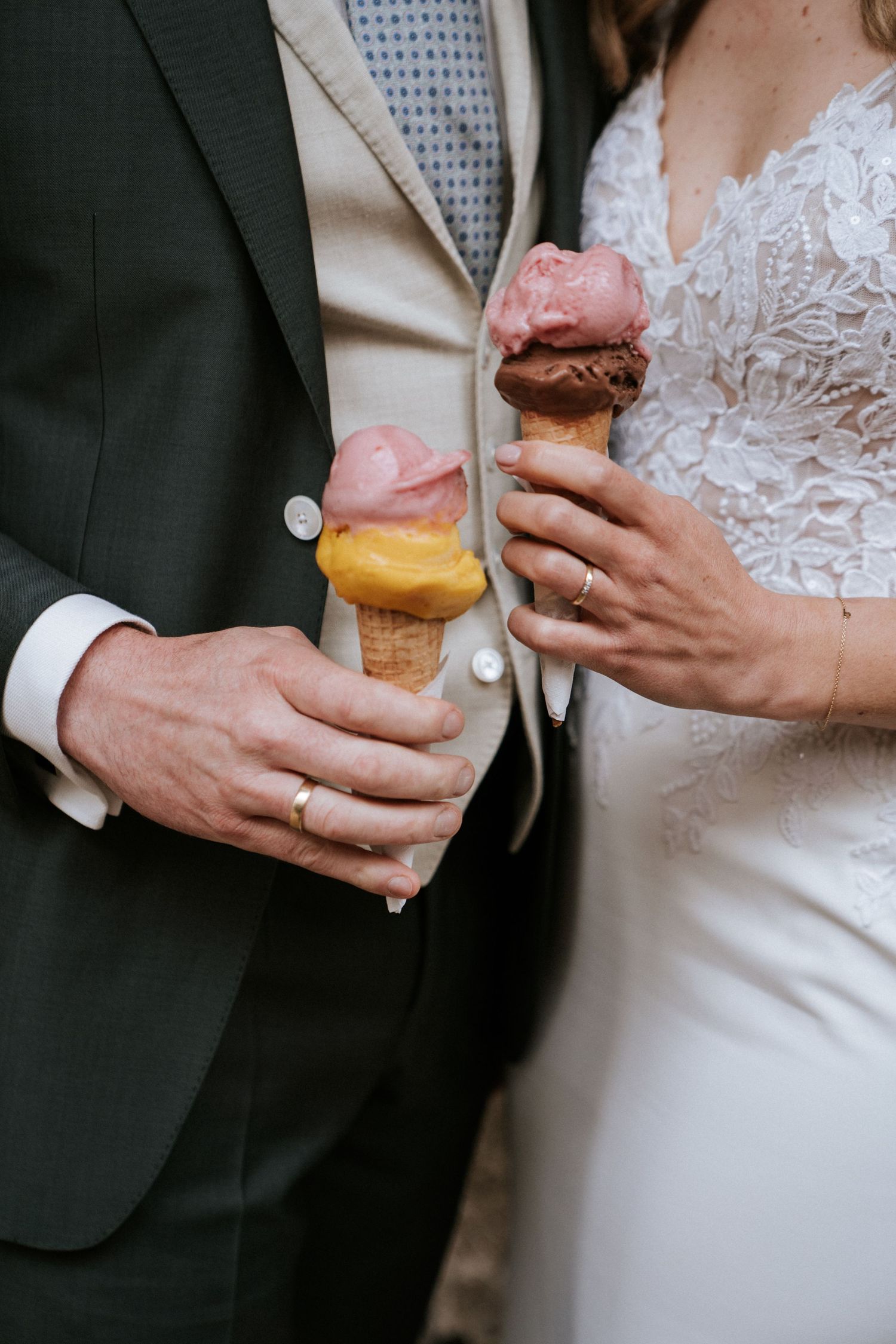 A couple in formal wedding attire holding pink ice cream cones, showing their hands with wedding rings.